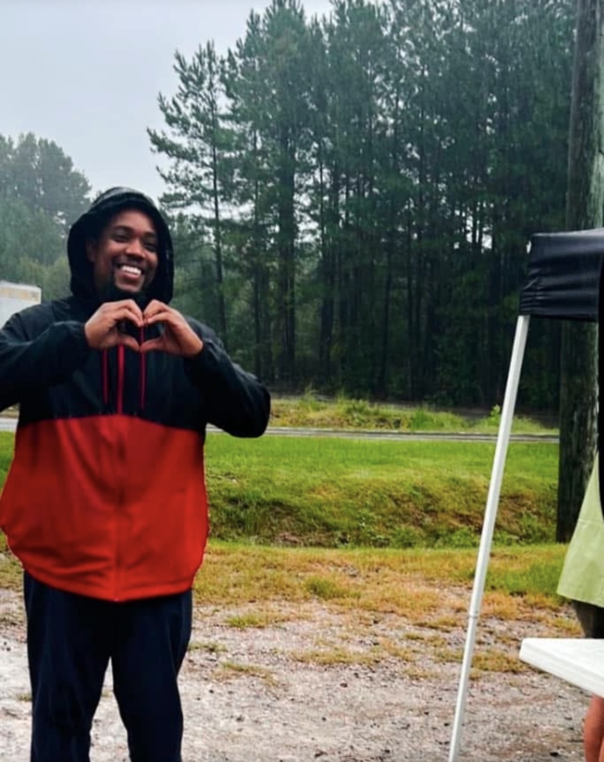 Person outdoors making a heart shape with hands, trees and grass in background.