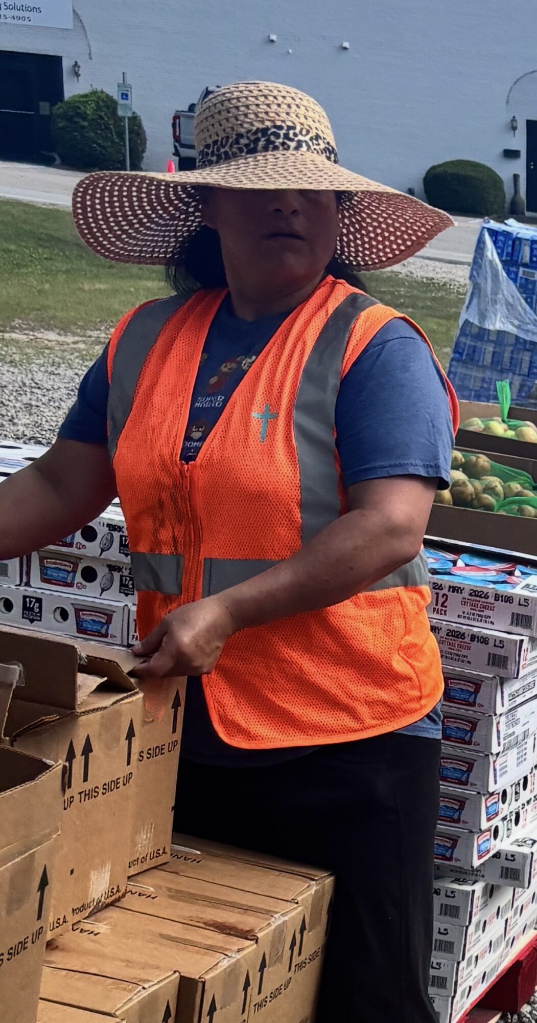 Person wearing a large straw hat and orange safety vest, standing outdoors near boxes and a cart.