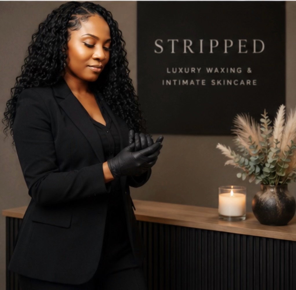 Woman with curly hair wearing black gloves and blazer, standing near a sign that reads 'STRIPPED' with skincare products.