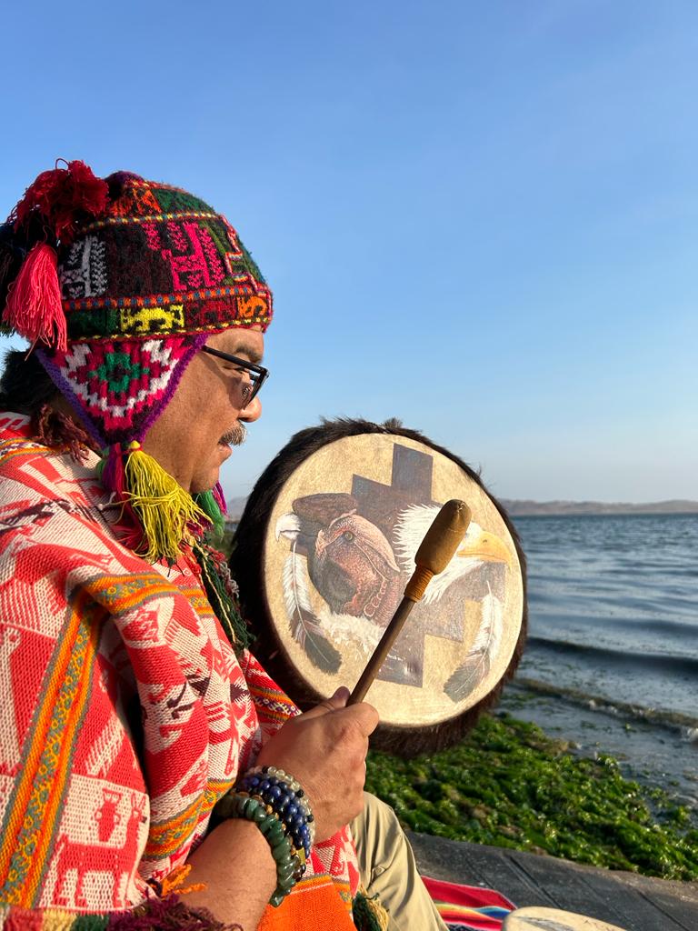 Woman wearing colorful clothing and a knit hat, playing a drum outdoors near water, with a clear sky in the background.
