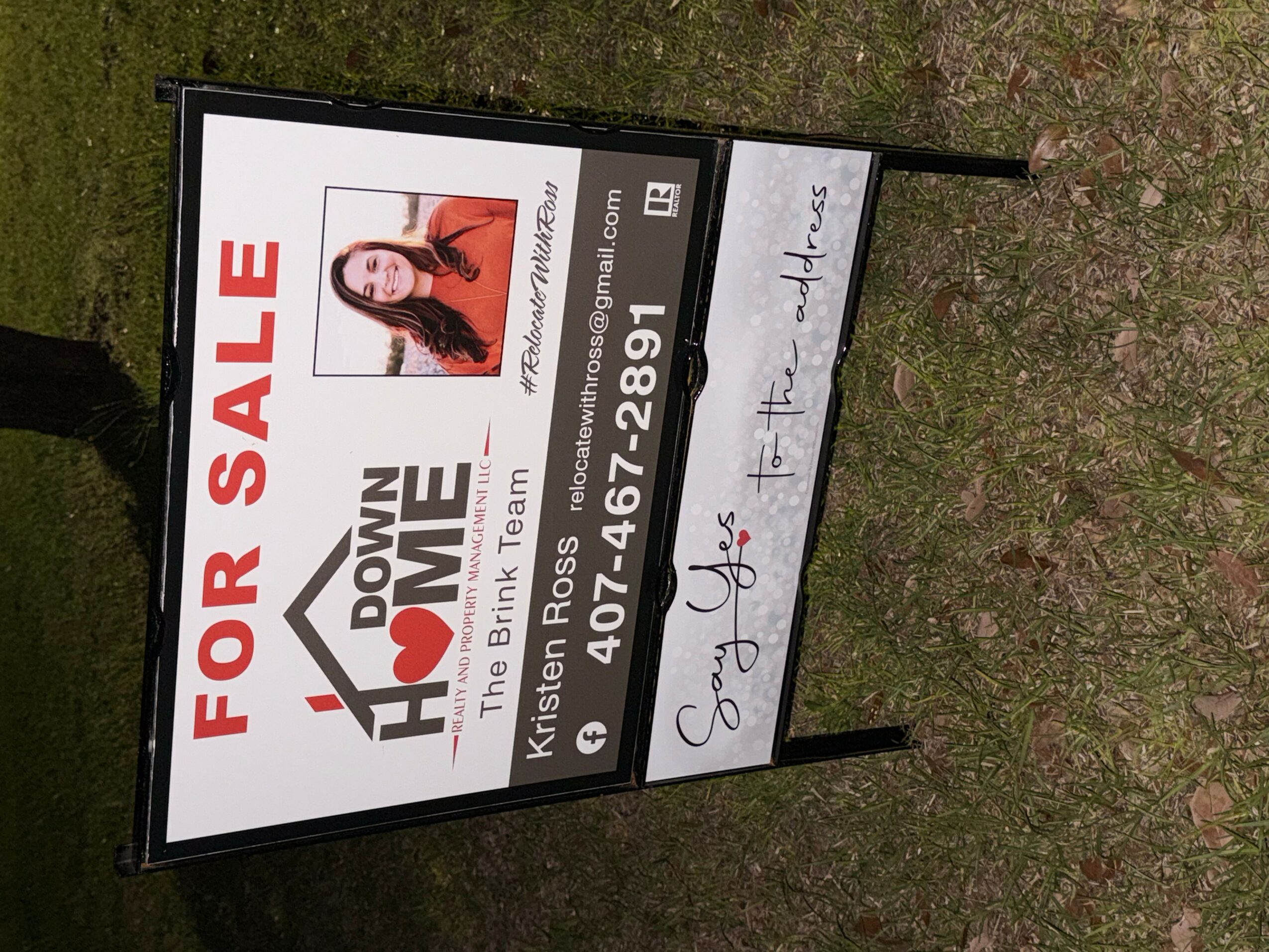 A sidewalk sign advertising a sale, with text and a photo of a woman, on grass and dirt ground.