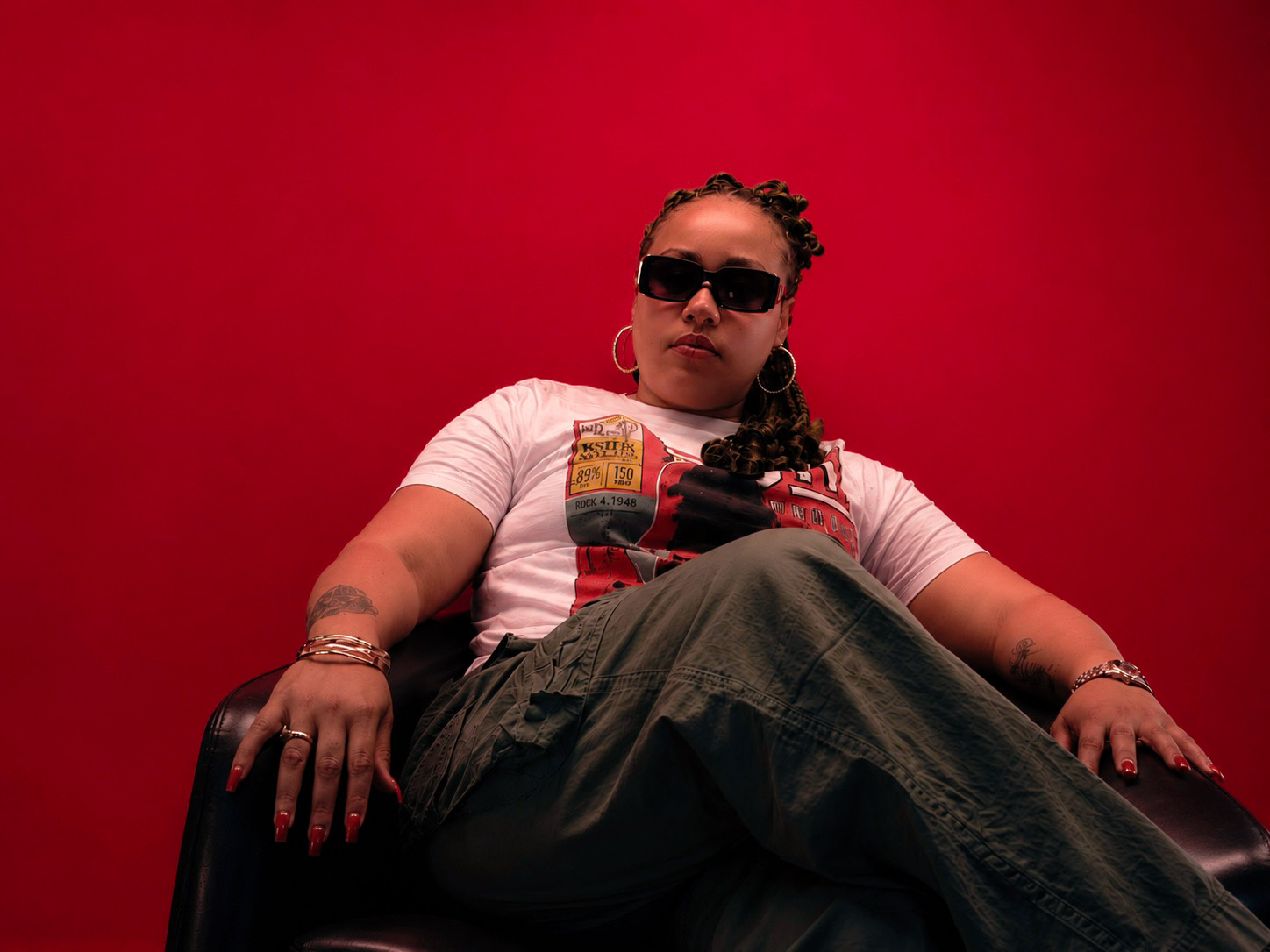 Woman sitting with crossed legs against a red background, wearing sunglasses, a white t-shirt, and dark pants.