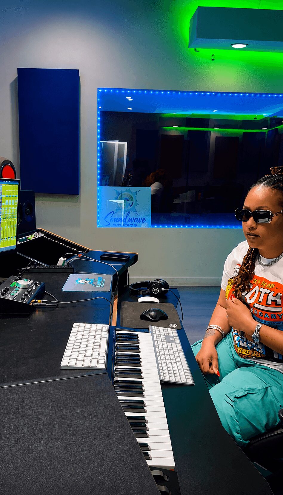 Person wearing sunglasses sitting in a music studio with keyboard, computer, and colorful lighting.