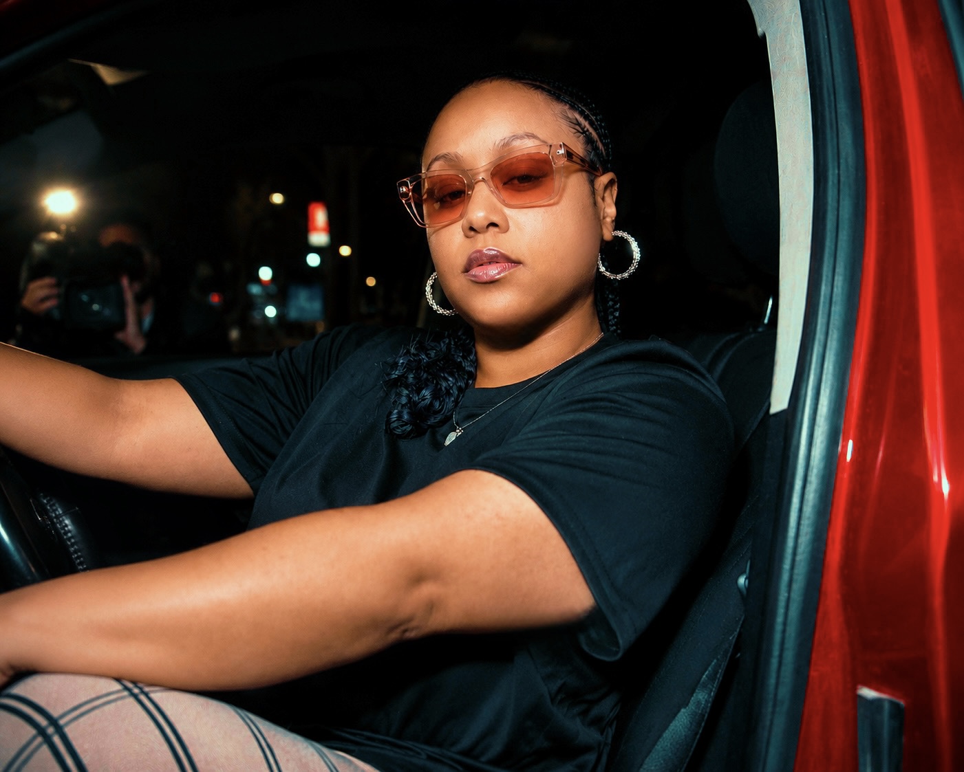 Woman with sunglasses and hoop earrings sitting in a vehicle at night, city lights in background.
