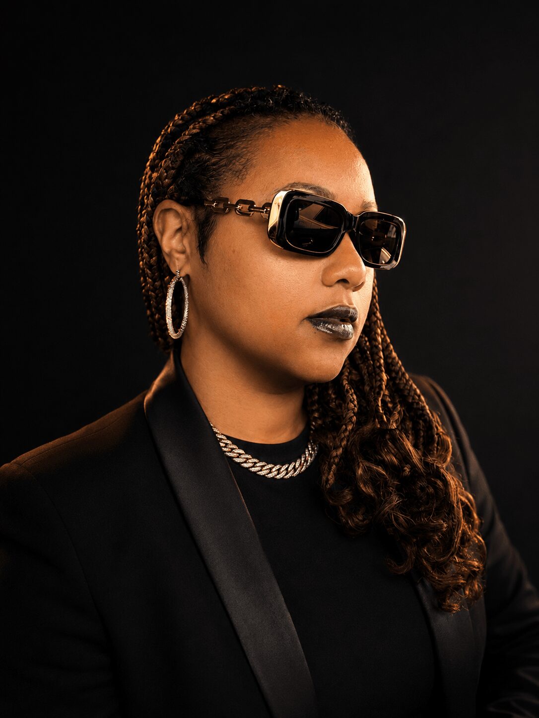 Woman with braided hair, wearing large sunglasses, hoop earrings, and dark lipstick, dressed in black against a dark background.