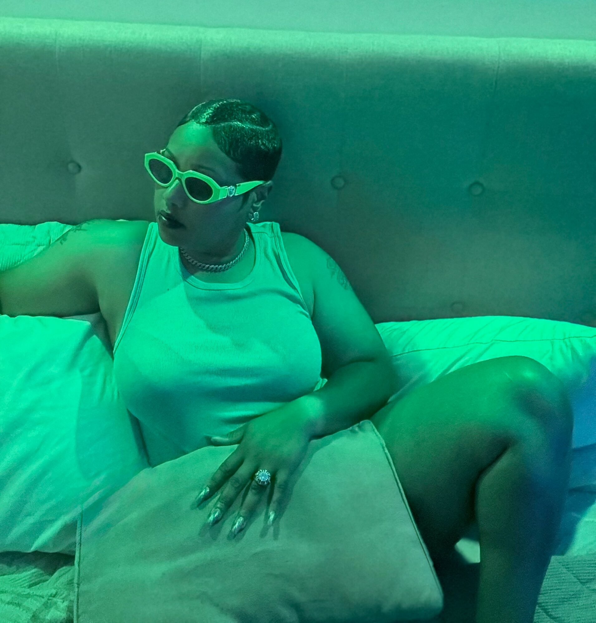 Person with short hair, wearing glasses, a sleeveless top, sitting on a bed with a pillow, in a greenish lighting.