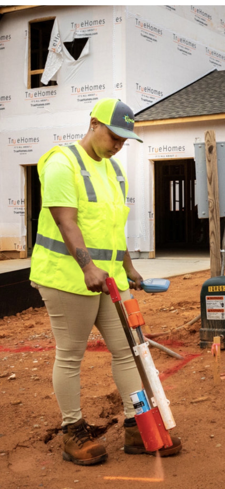 Construction worker in high-visibility clothing and helmet using a tool on dirt site, partially built house in background.