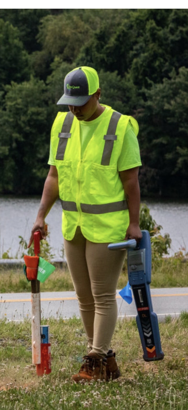 Person in high-visibility vest and helmet holding a measuring tool outdoors near water, trees in background.