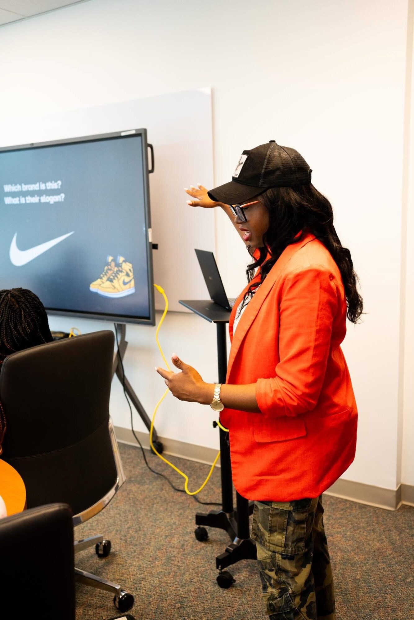 Woman in orange jacket and camouflage pants using a smartphone in a room with a large screen displaying Nike logo and shoes, black chair, and white wall.