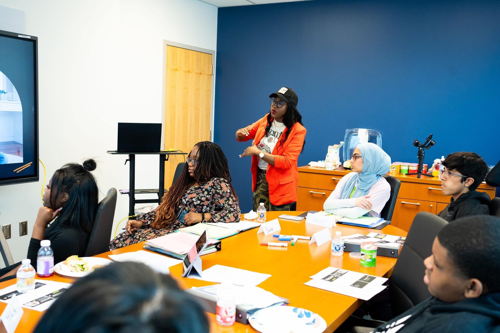 Group of people in a meeting room watching a presentation on a screen, with a woman standing and speaking.