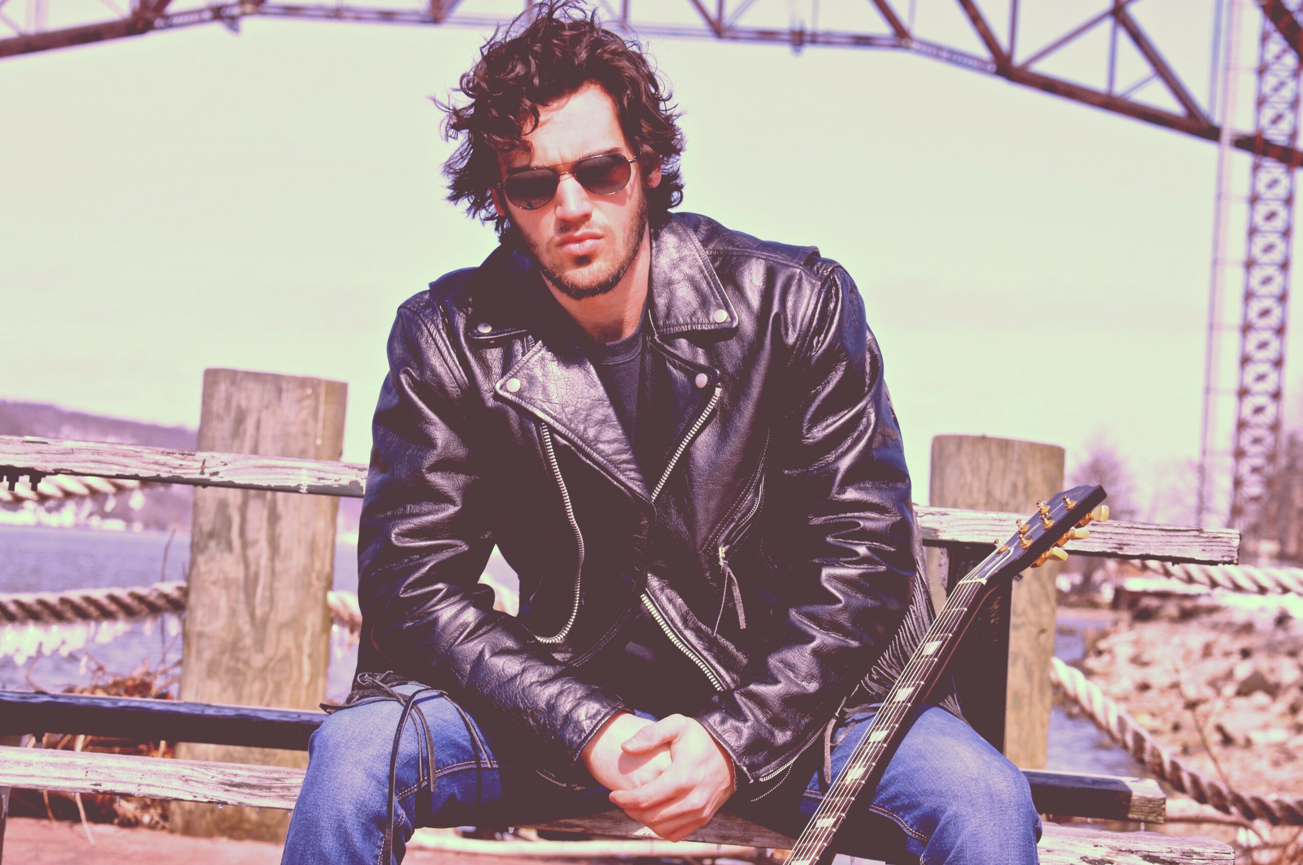 Person with curly hair wearing sunglasses and a leather jacket sitting outdoors with a guitar, wooden posts, and a metal structure in background.