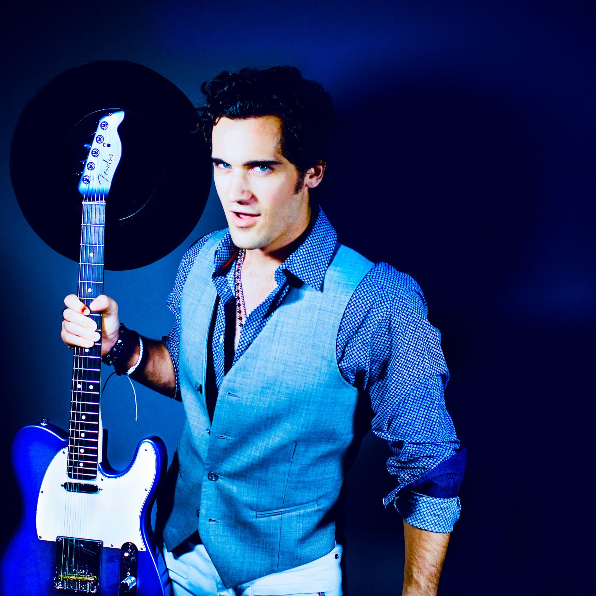 Young man with dark curly hair holding a white electric guitar, wearing a blue shirt and vest, against a dark background.