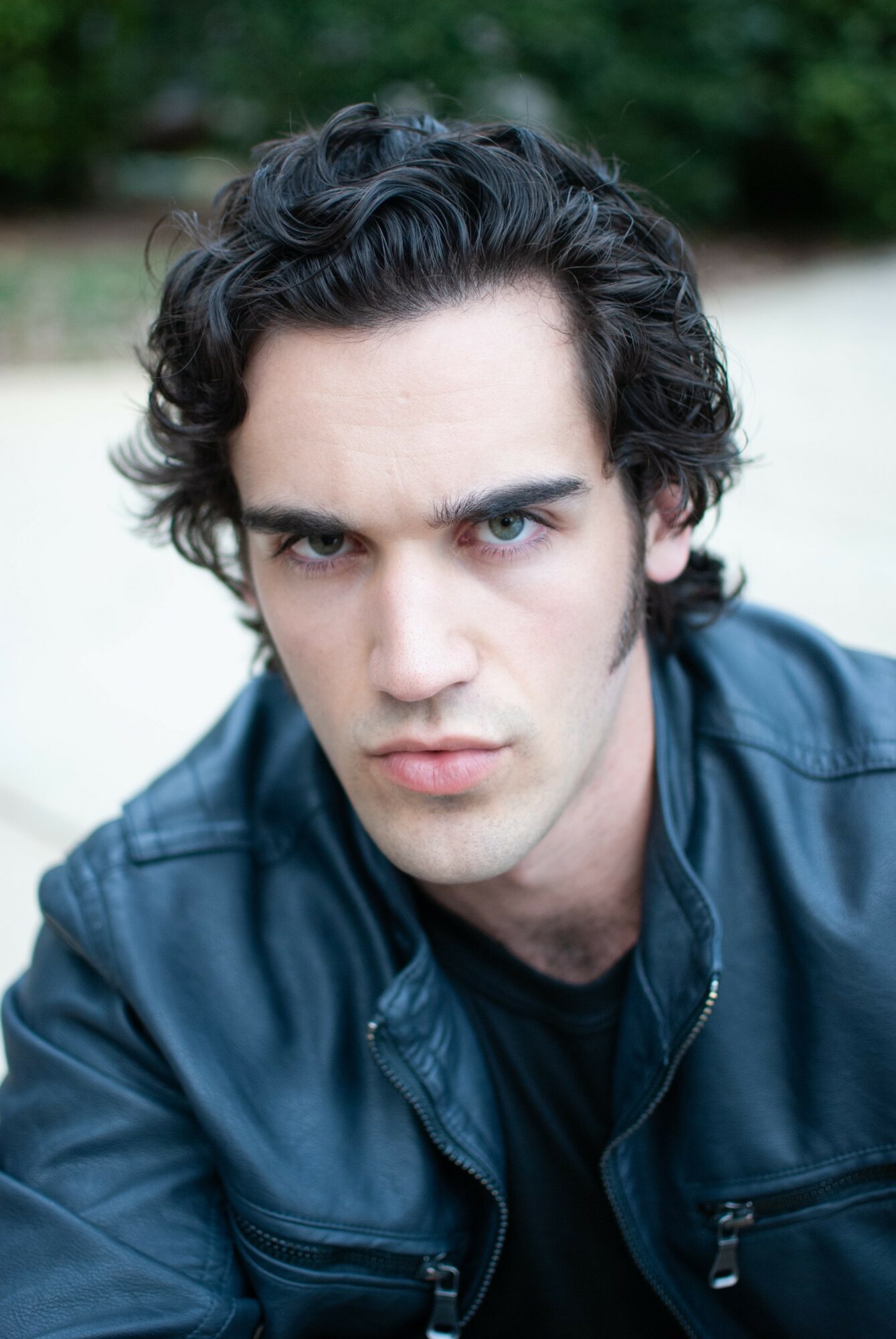 Young man with dark curly hair and intense gaze, wearing a dark jacket outdoors, with blurred greenery in background.