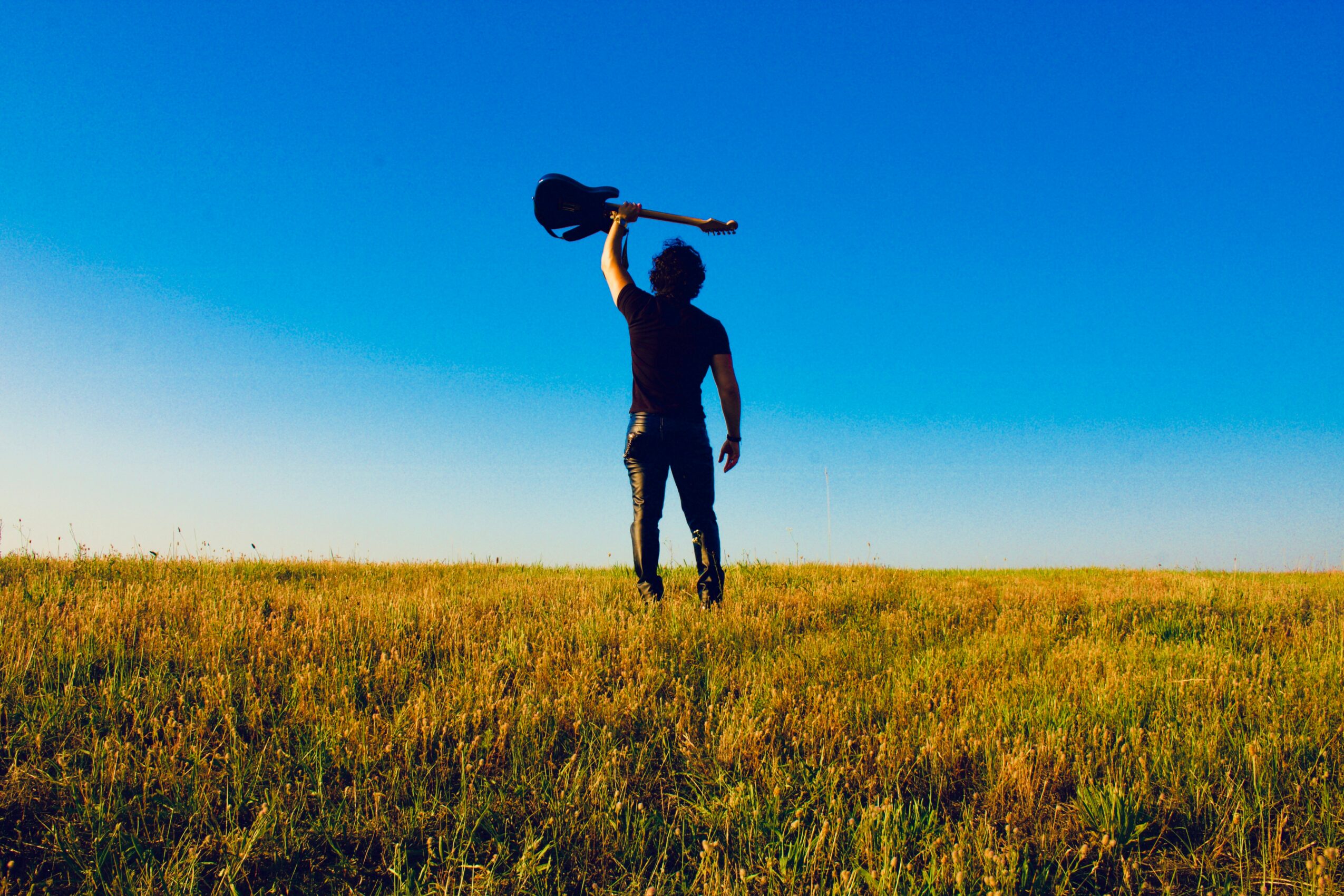 Person standing in a grassy field holding a guitar over their shoulder against a clear blue sky.