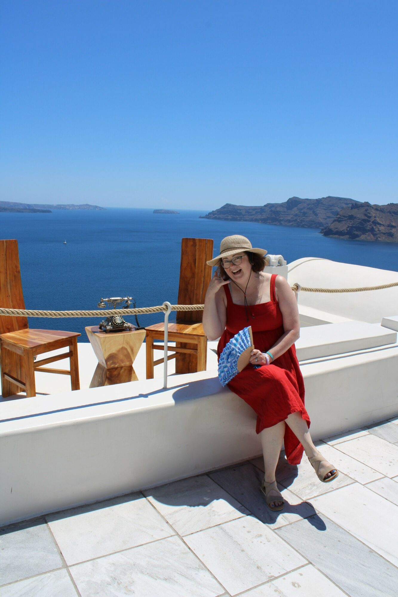 Person in red dress sitting on a white ledge near water with mountains in the background, holding a bag, sunny day.