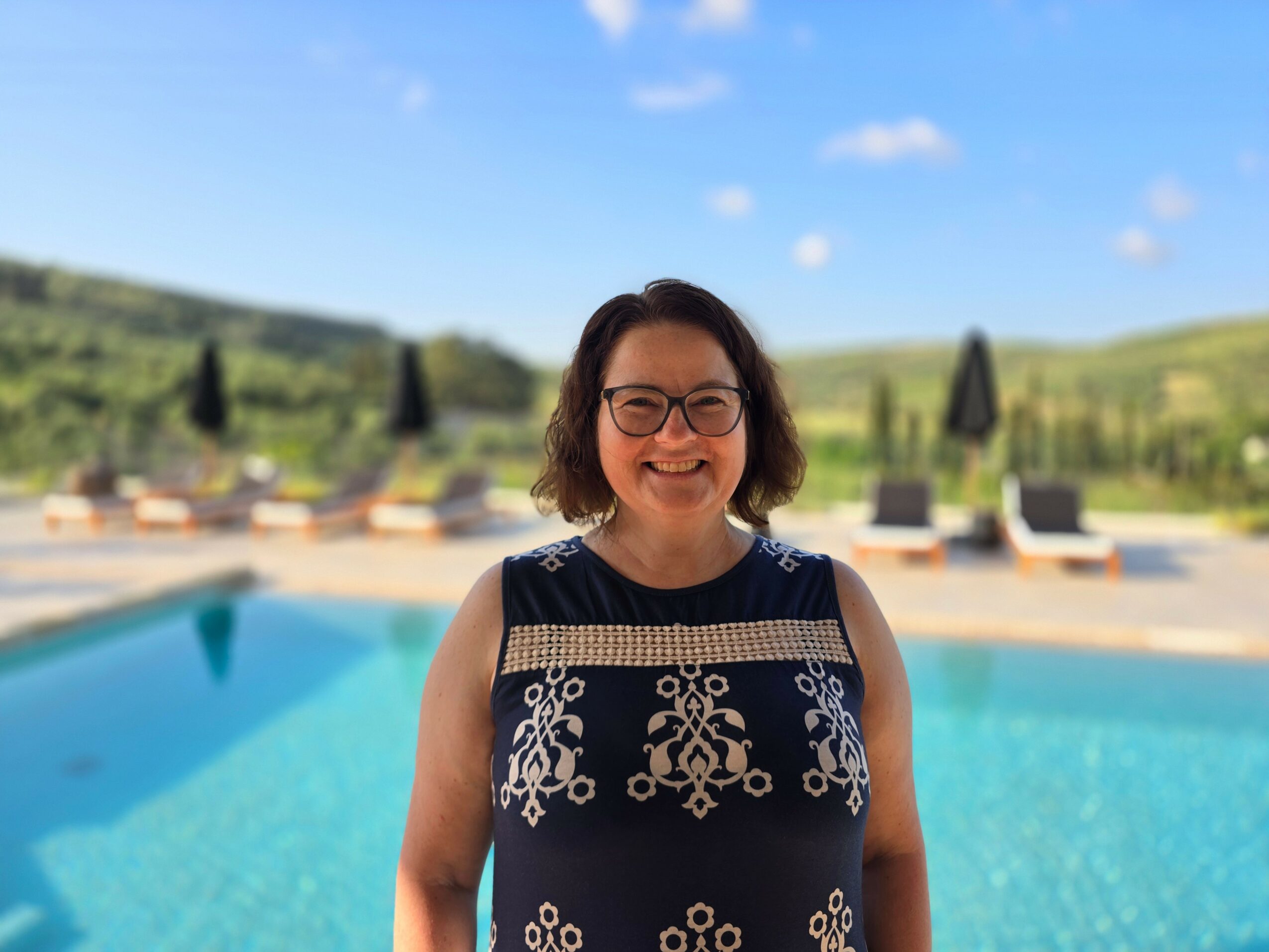 Smiling woman with glasses and shoulder-length hair standing by a swimming pool outdoors, with a landscape and blue sky in the background.
