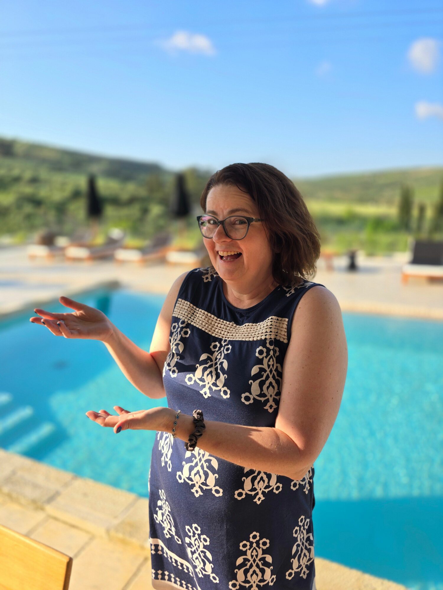 Woman with glasses in a patterned dress near a swimming pool, with a landscape background.
