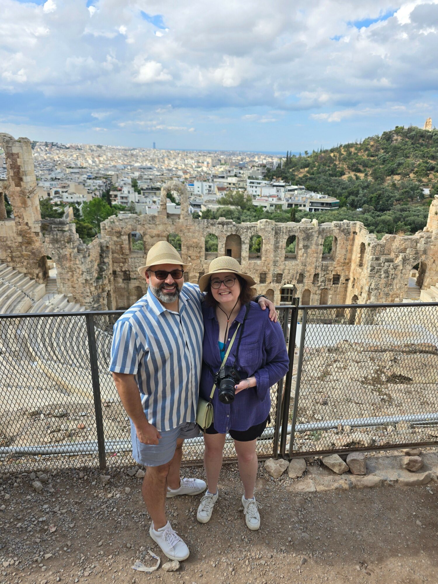 Two people stand outdoors near ancient ruins with a cityscape and cloudy sky in the background.