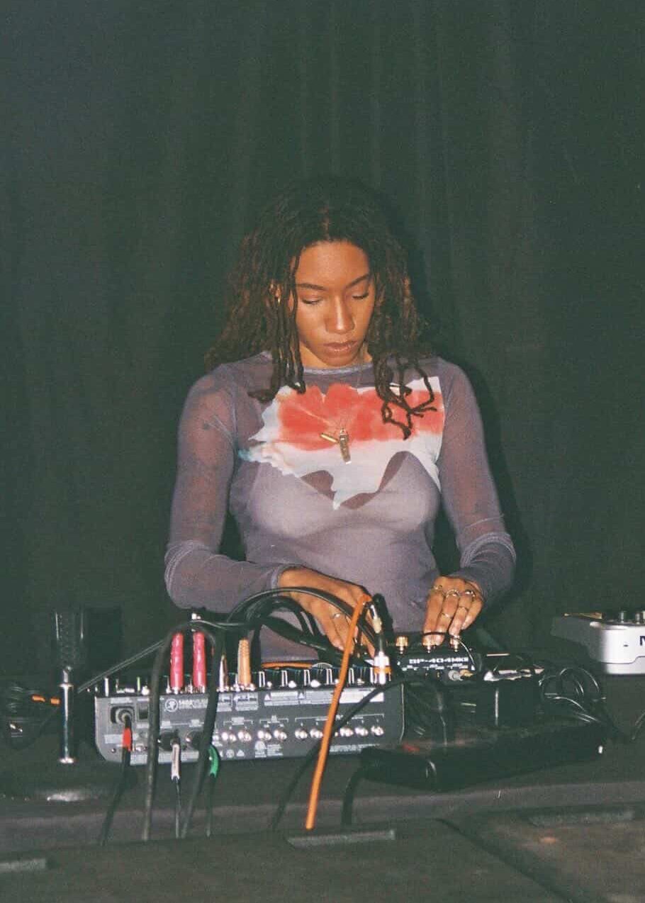 Woman with curly hair operating DJ equipment on stage, dark background, focused expression.