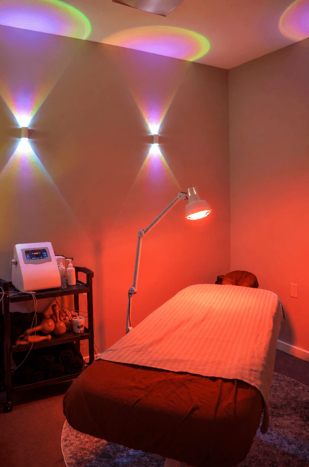 A massage or treatment room with colorful lighting, a bed with a blanket, a lamp, and a small cart with items.