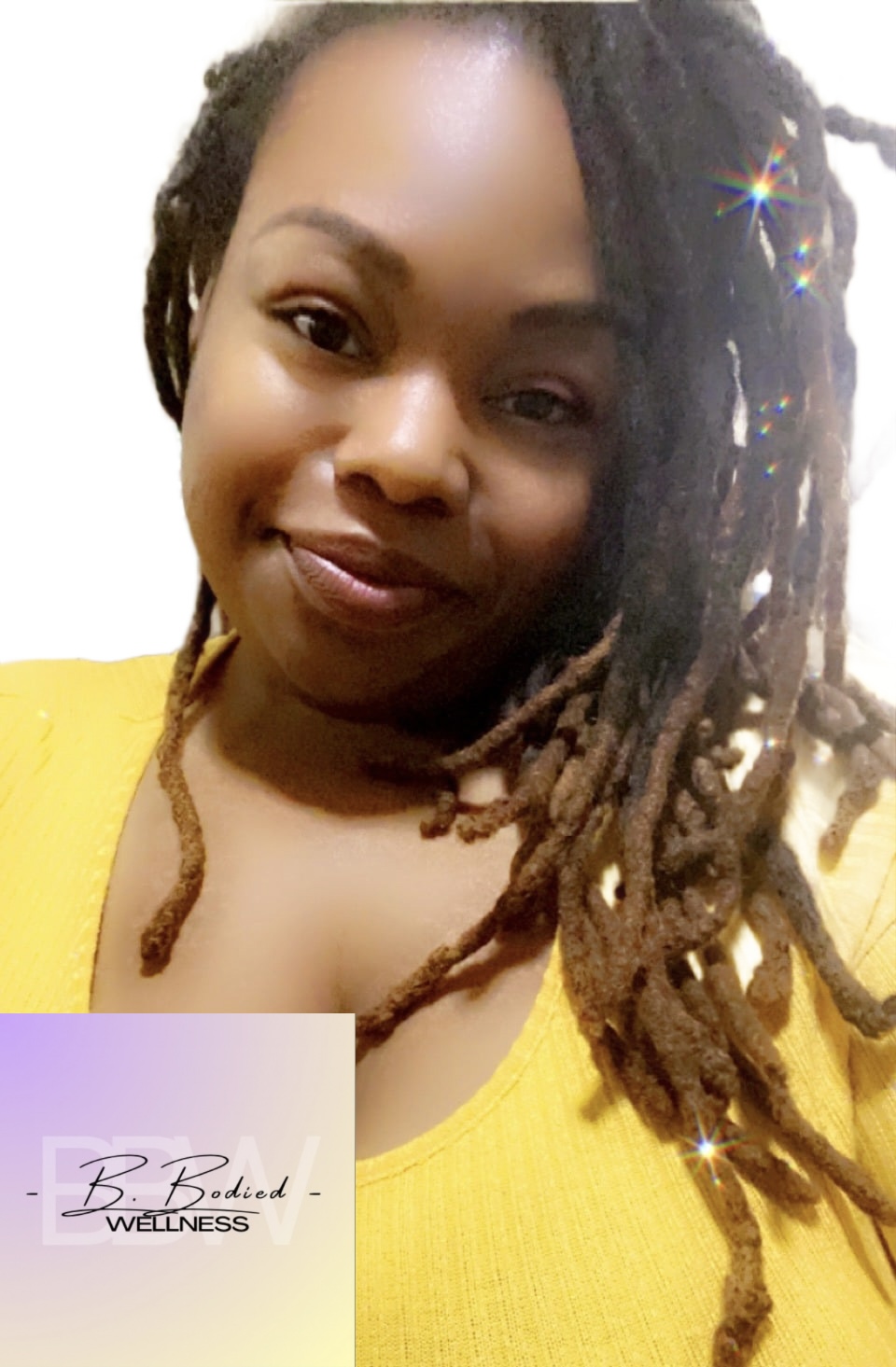 Woman with long dreadlocks wearing a yellow top, smiling at the camera, with a light background.