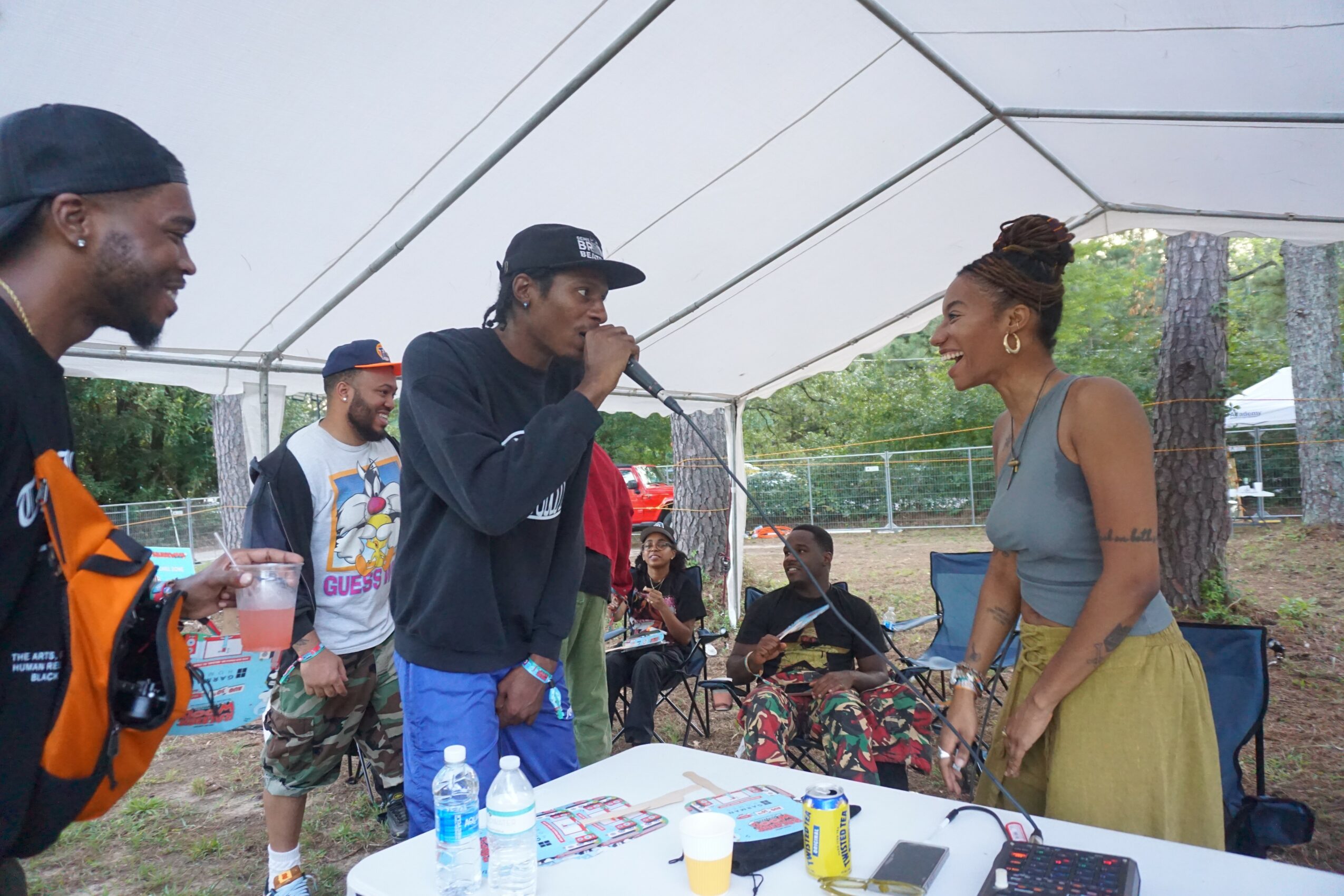 Group of people outdoors under a tent, woman smiling at man holding a microphone, table with drinks and items, trees in background.