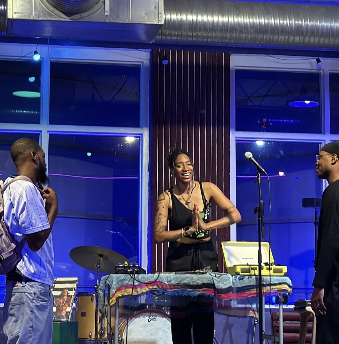Three people on stage with musical equipment, smiling and talking, in a blue-lit room with large windows.