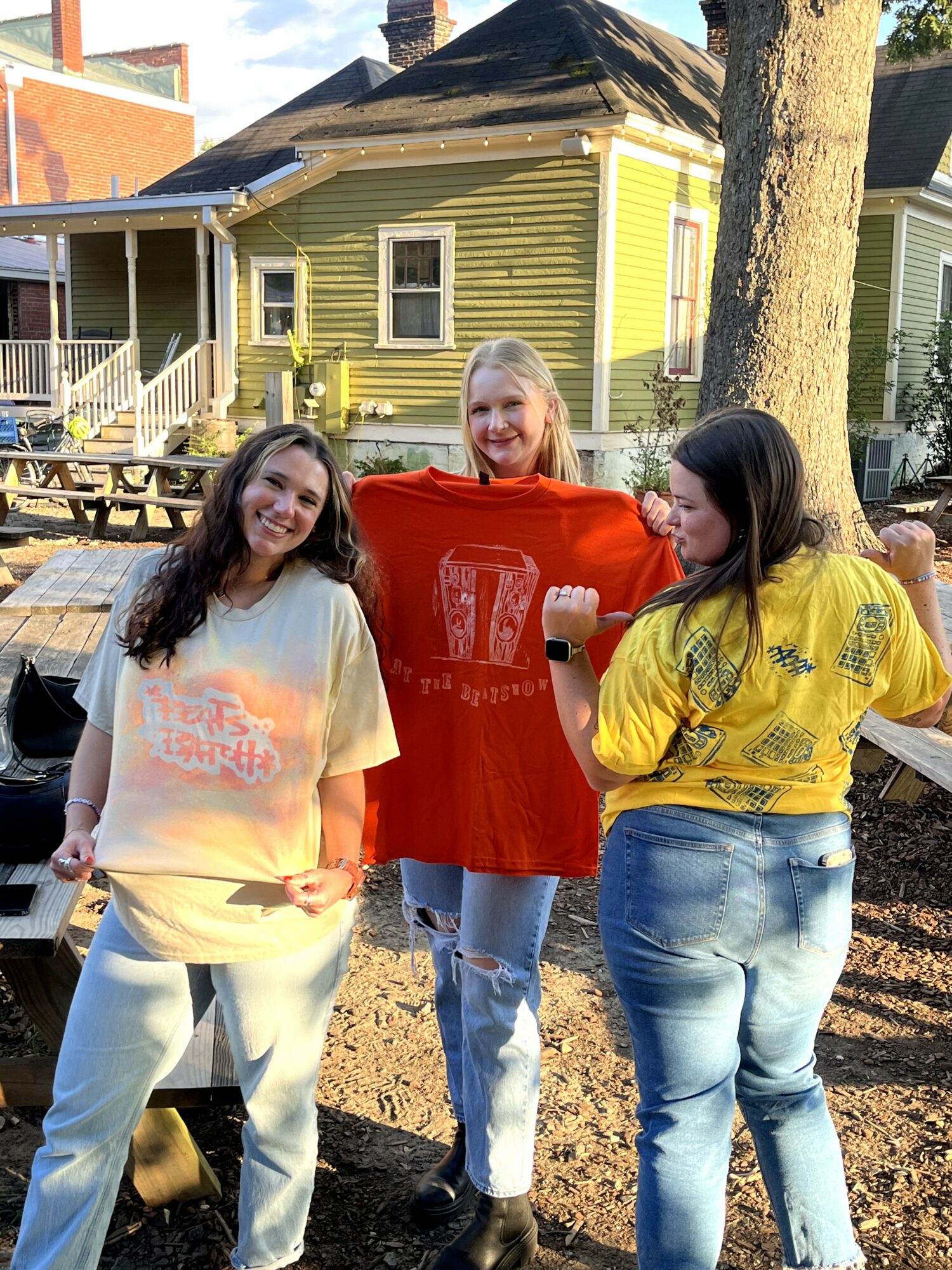 Three women standing outdoors in front of a house, smiling, with trees and a picnic table nearby.