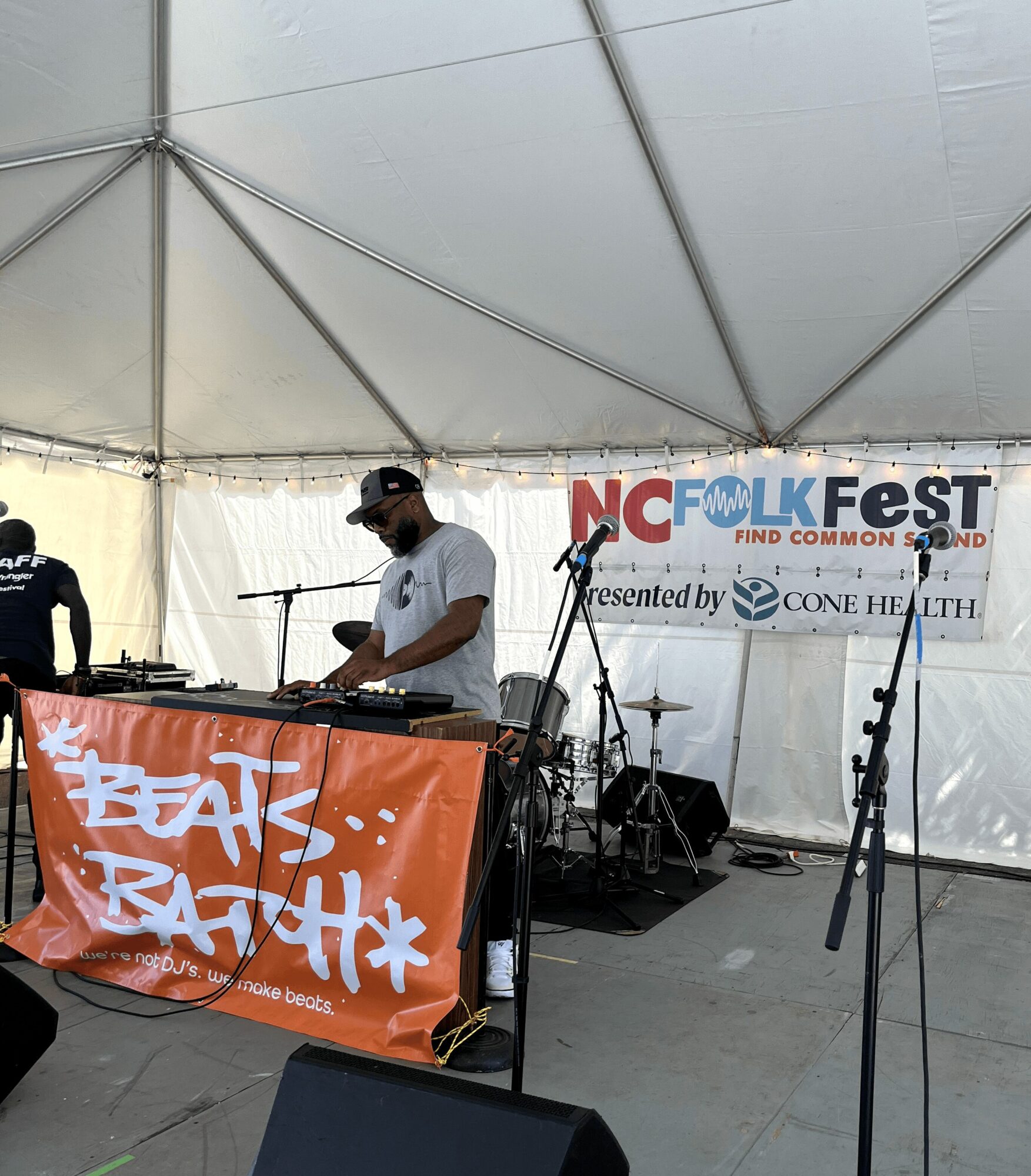 A person wearing a cap and sunglasses stands behind a DJ setup under a tent, with a banner reading 'NC Folk Fest' in the background.