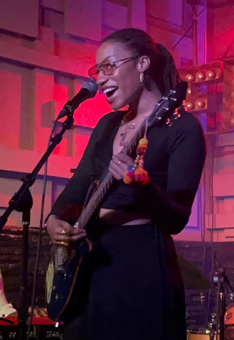 Woman singing into microphone while playing guitar on stage with colorful background, smiling, wearing glasses and a dark outfit.