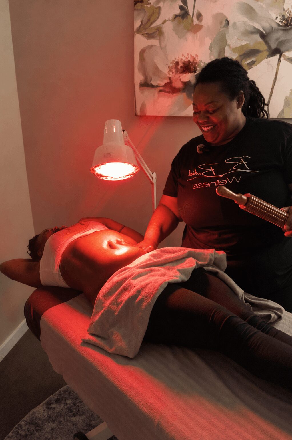 A woman smiling and holding a tool next to a person lying on a massage table with a towel, under a red light.