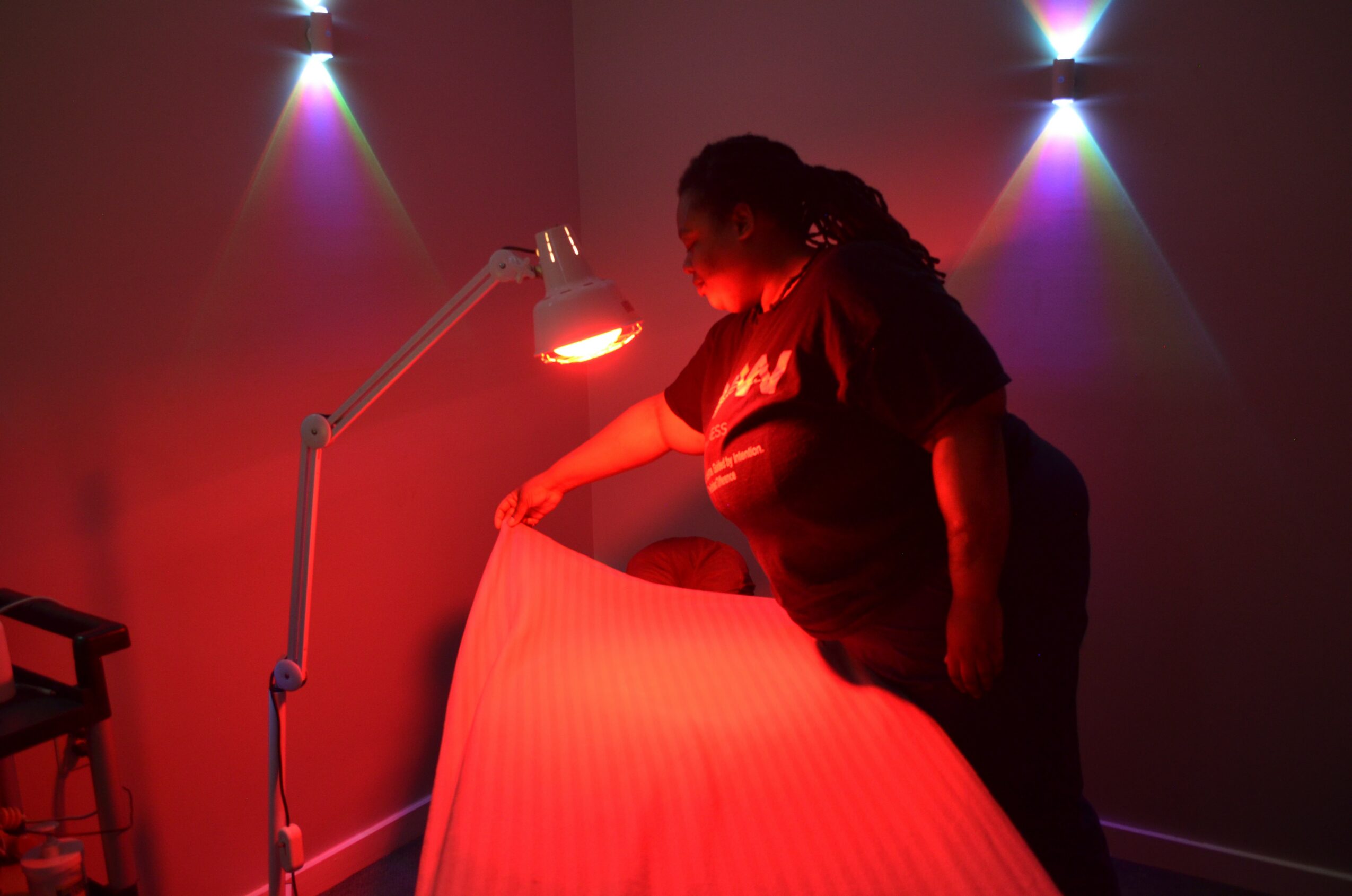Person adjusting a bed under a red light in a dimly lit room with colorful wall lights, holding a sheet or blanket.