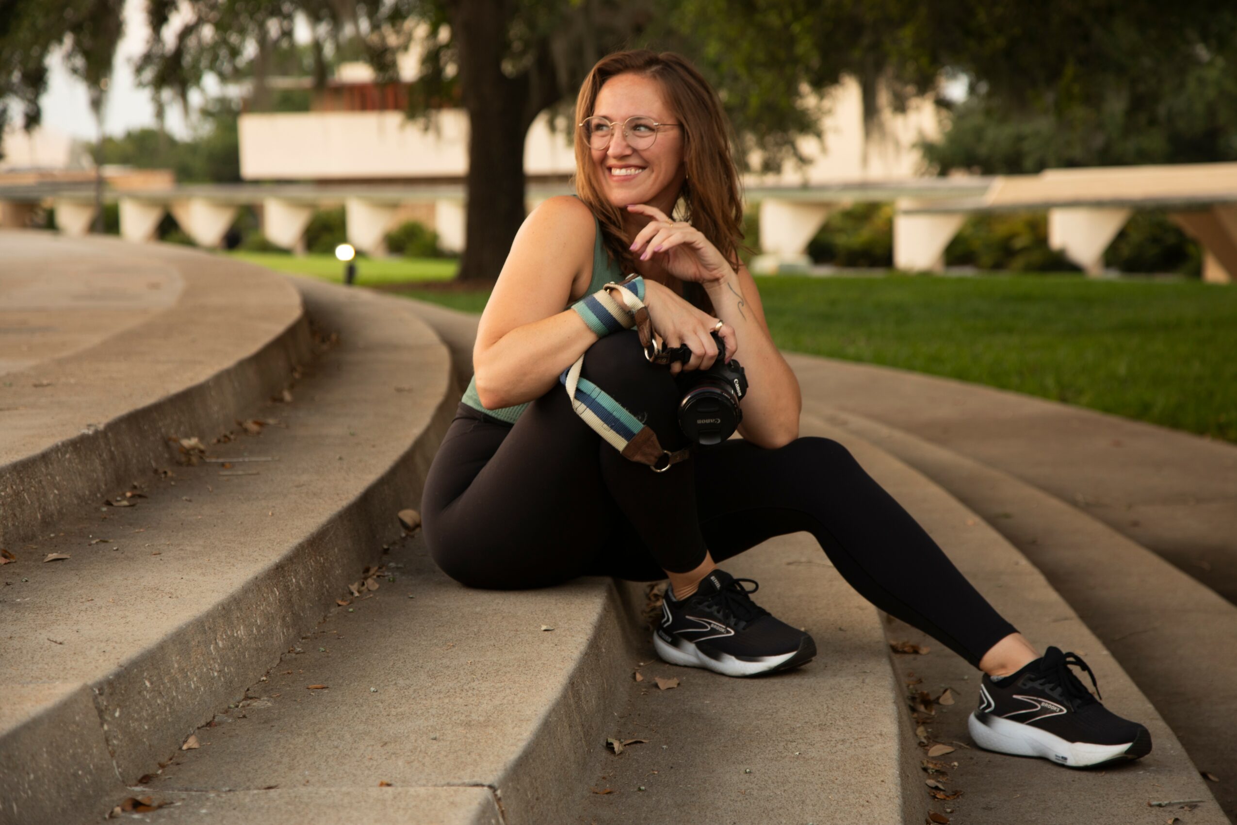 Woman sitting on steps outdoors, smiling, holding a camera, with trees and buildings in background.