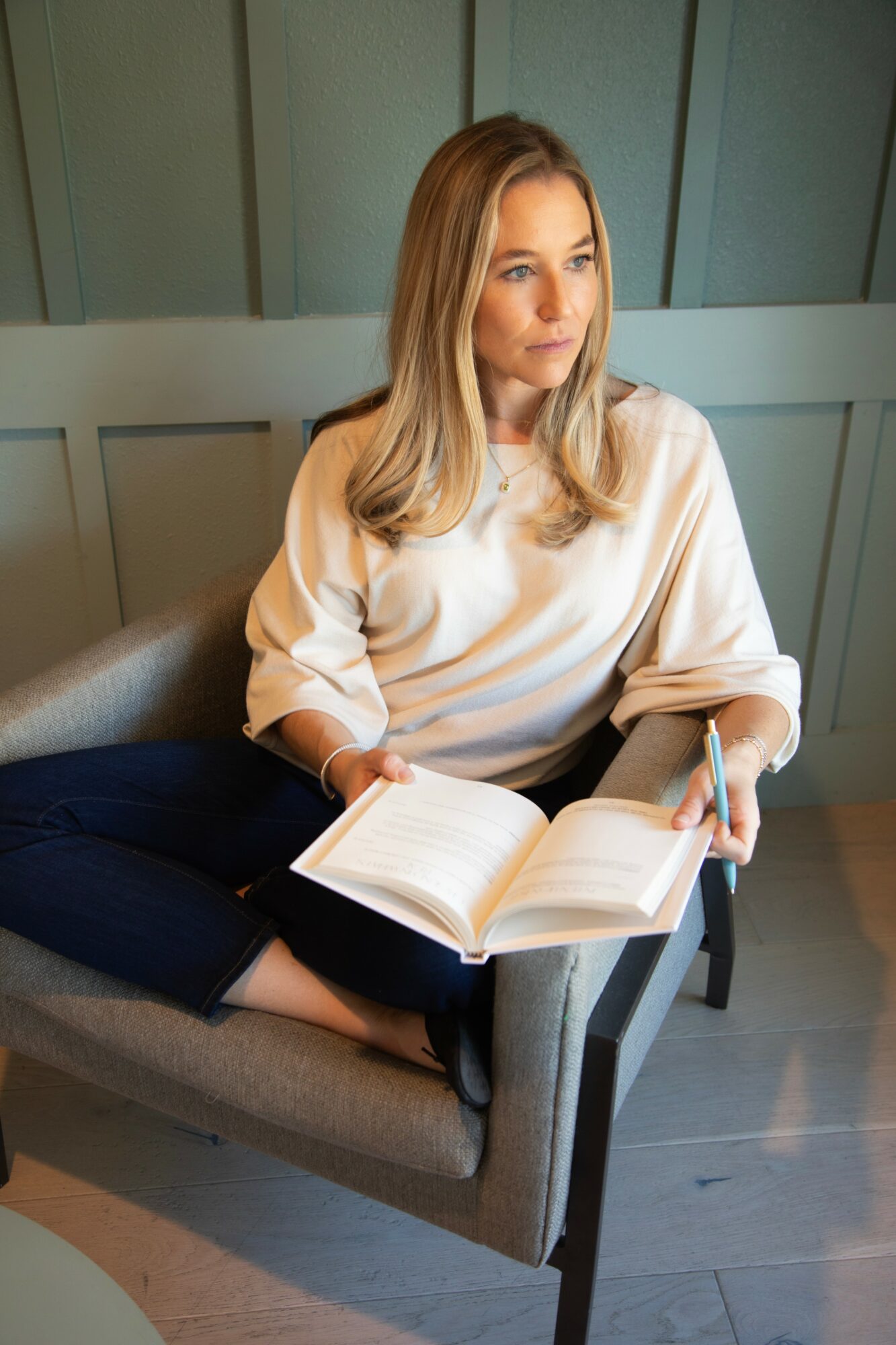Woman with long blonde hair sitting on a chair, holding an open book, looking to the side, in a room with green walls.