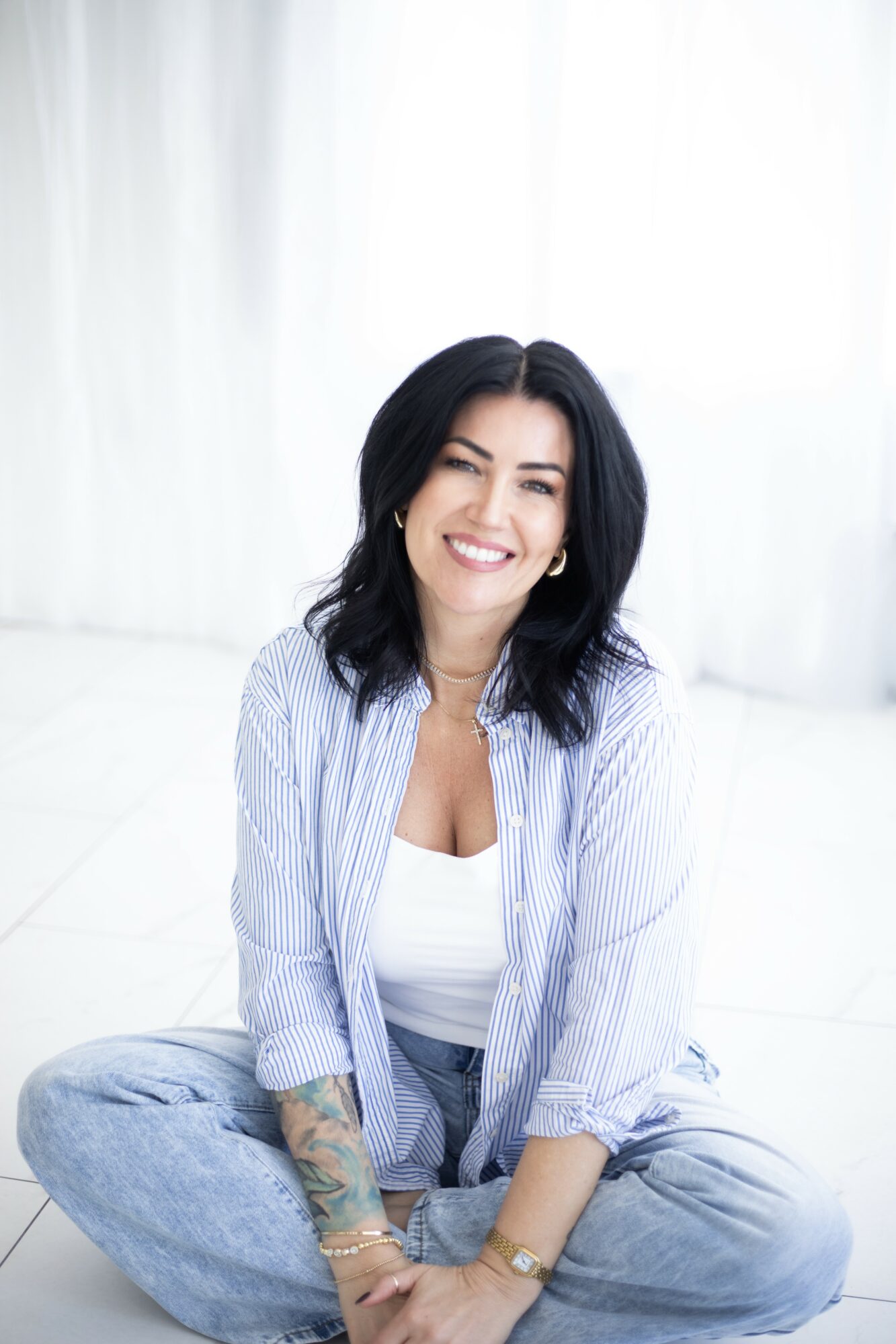Smiling woman with dark hair sitting cross-legged on the floor, wearing a striped shirt and jeans, in a bright room.