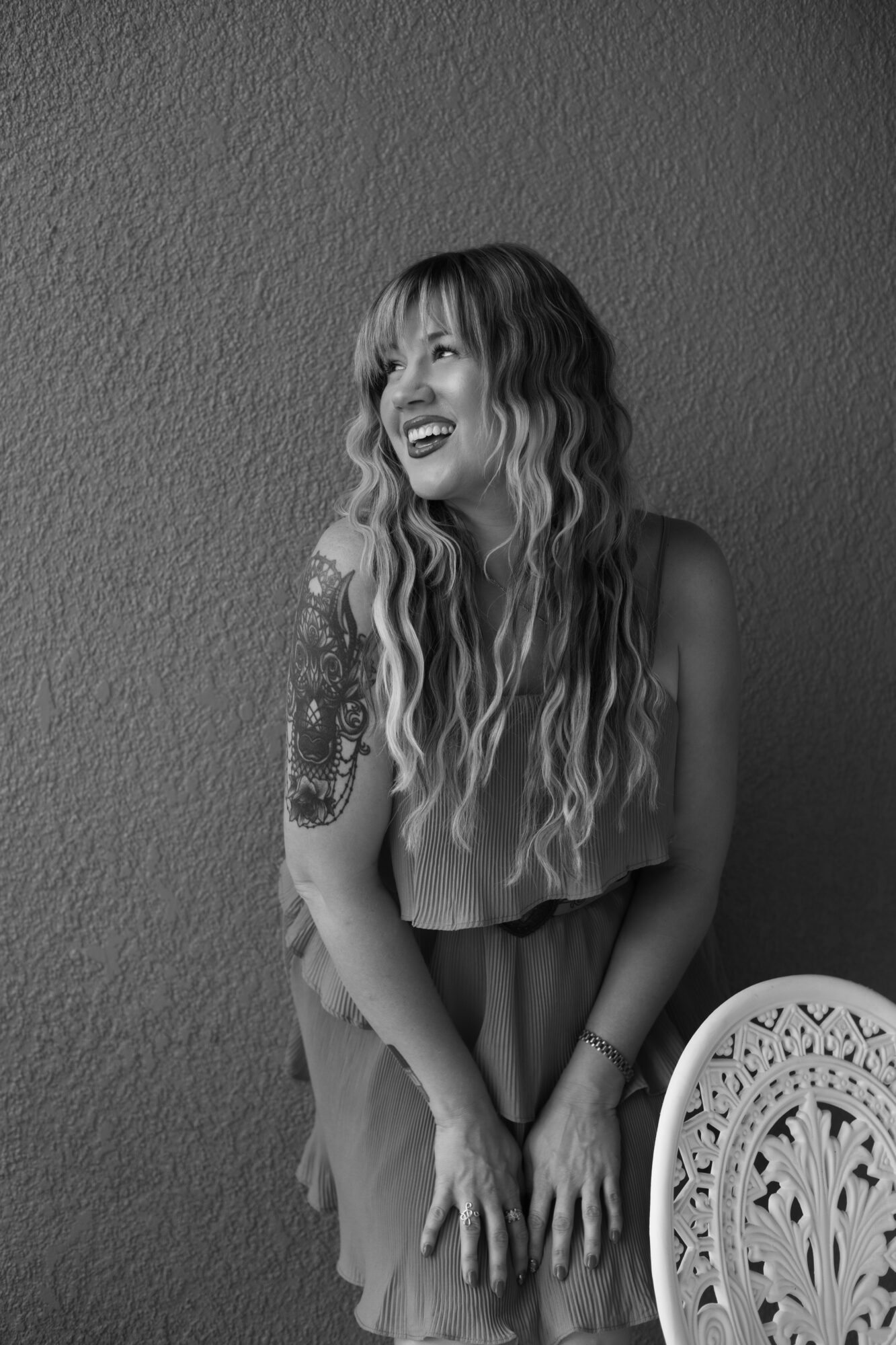 Woman with long wavy hair smiling, standing against a textured wall, holding her hands in front, with a decorative chair nearby.