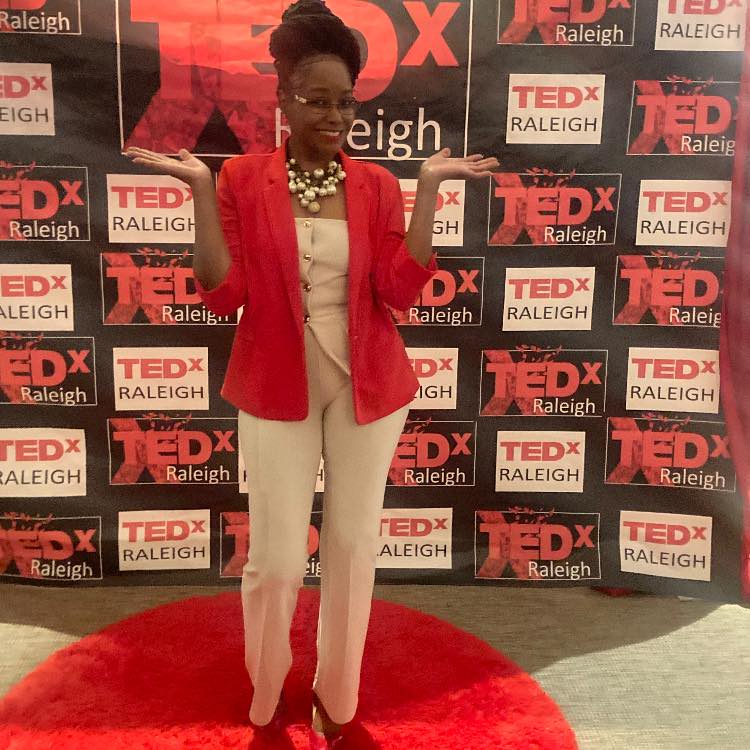 Woman in red blazer and white pants standing on red carpet in front of TEDx Raleigh backdrop.