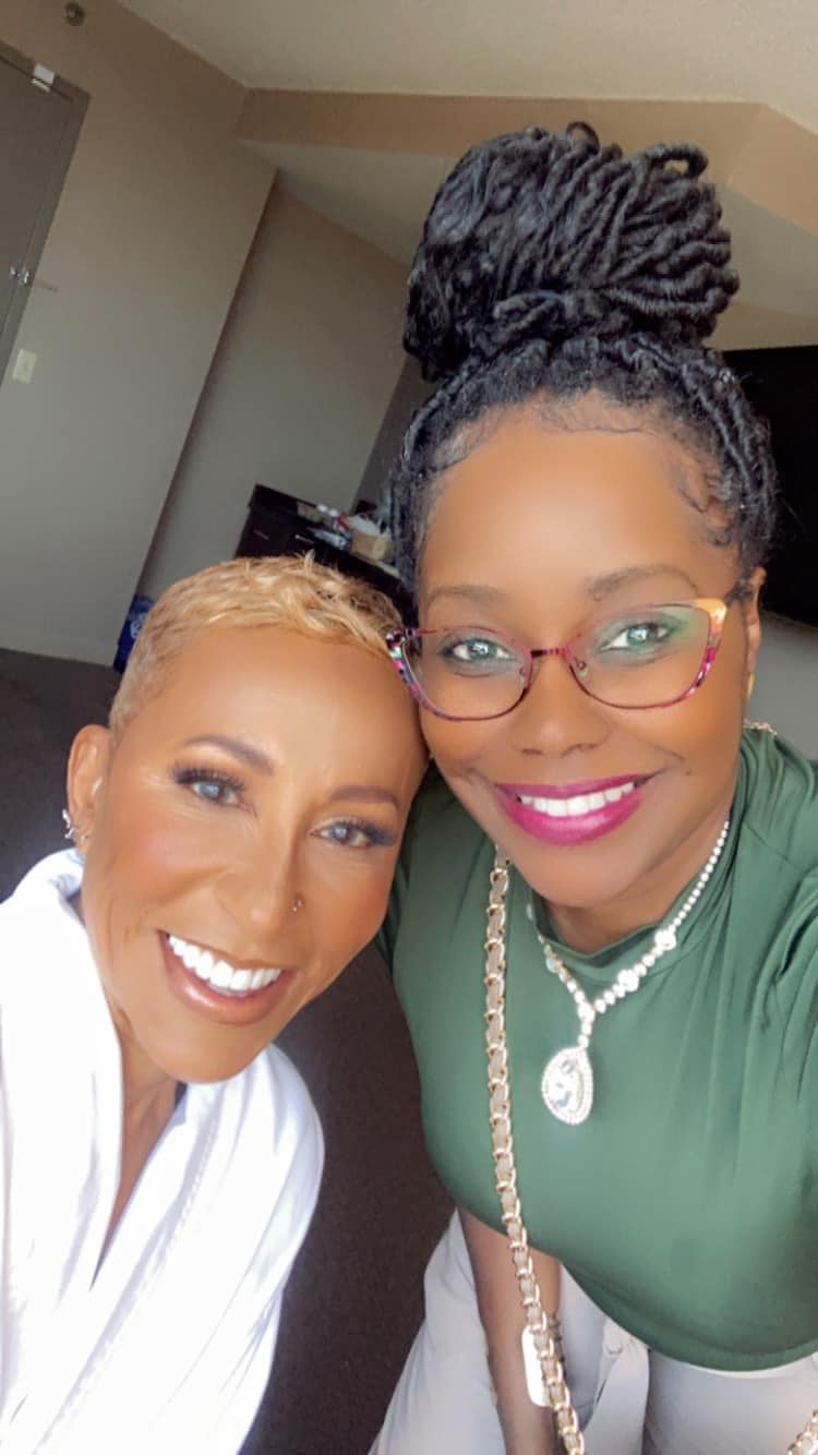 Two women smiling, one with glasses and a green top, the other with short hair and a white shirt, indoors.