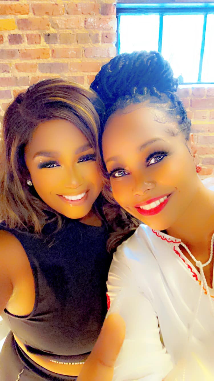 Two women smiling closely, one with dark hair and the other with braided hair, in front of a brick wall and window.