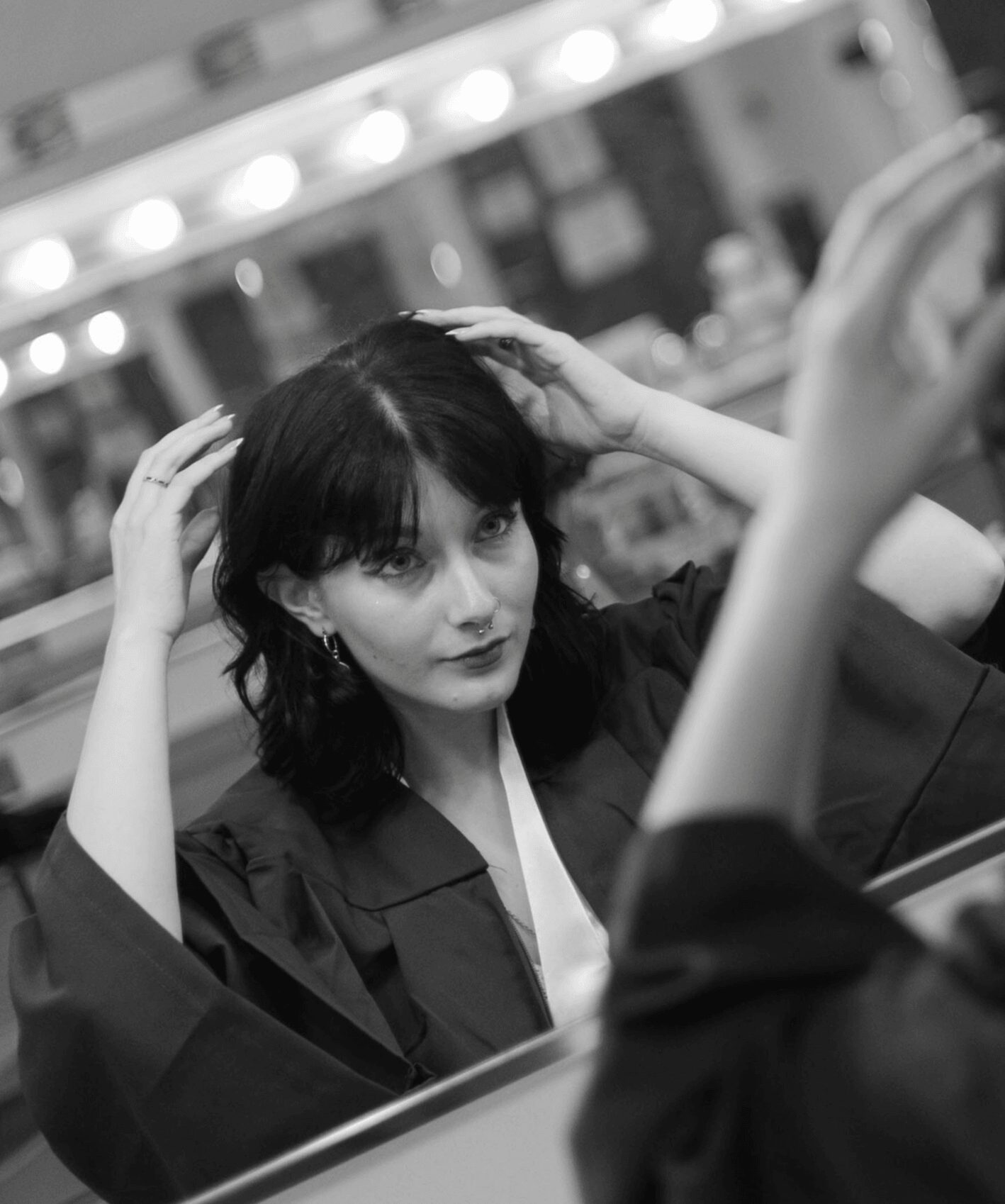 Woman with dark hair looking at mirror, adjusting her hair, in a dressing room with bright lights.