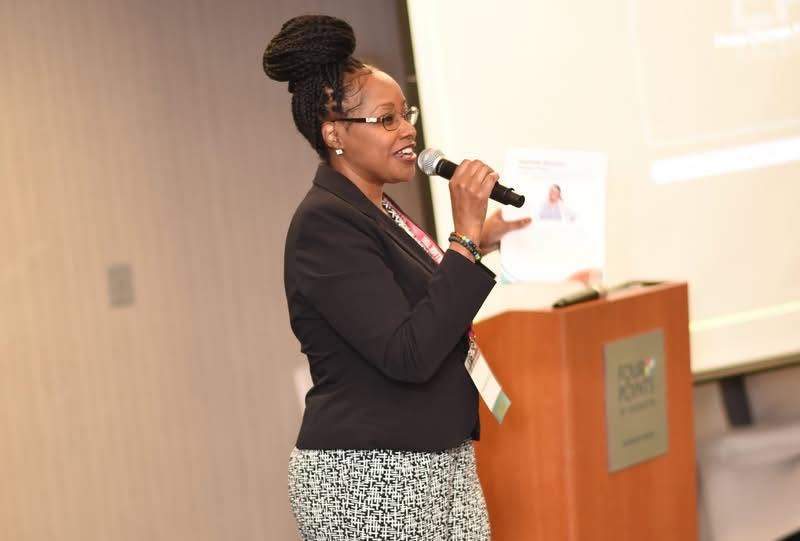 Woman speaking into a microphone at a presentation, standing next to a podium and a projection screen.