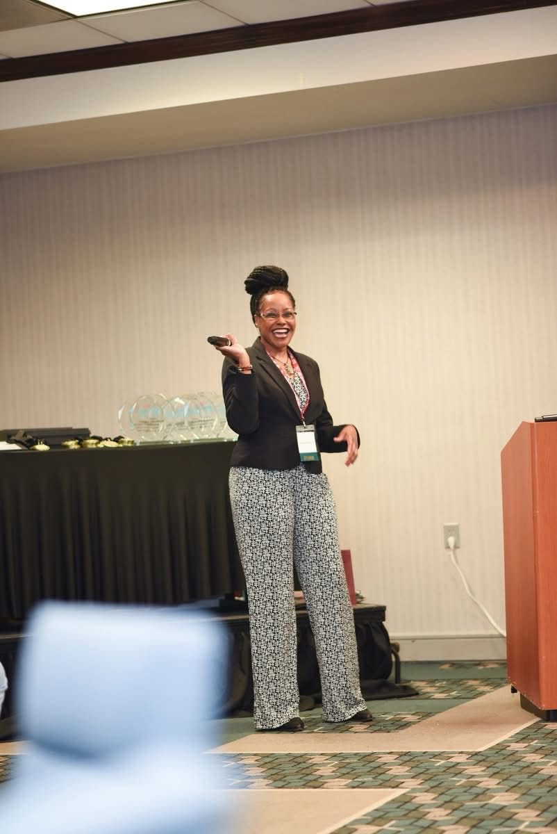 Woman standing and smiling in a conference room, holding a cup, with a table and a podium nearby.
