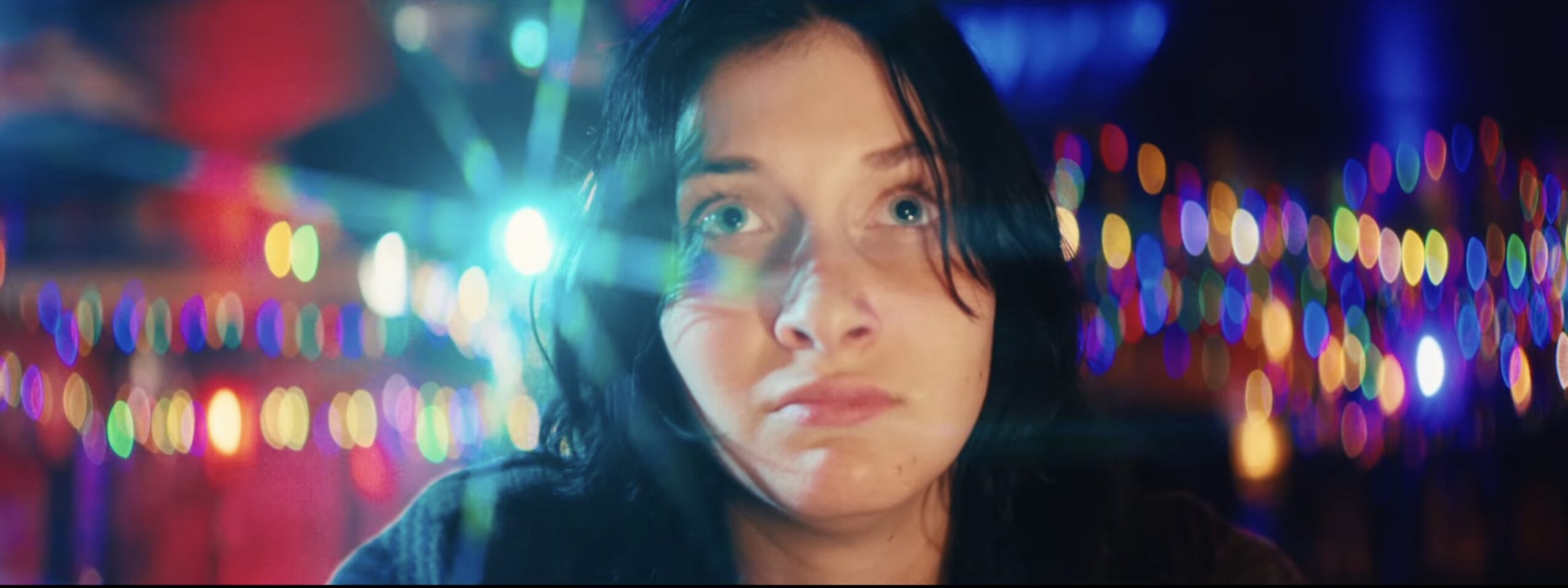 Close-up of a young woman with dark hair in front of colorful, blurred lights at night.