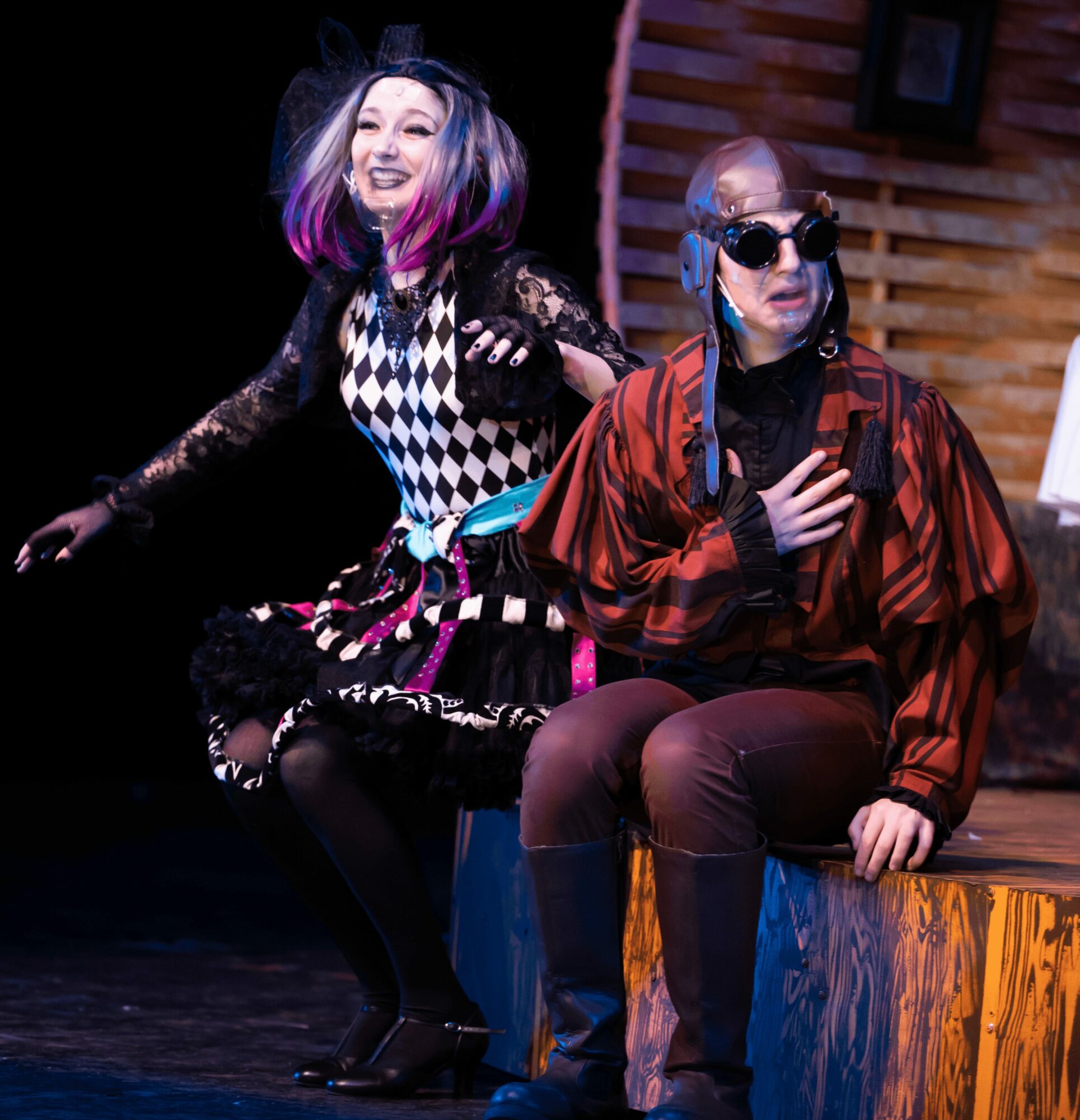 Two women sitting on a stage, one smiling with colorful hair, the other wearing sunglasses and a striped jacket.