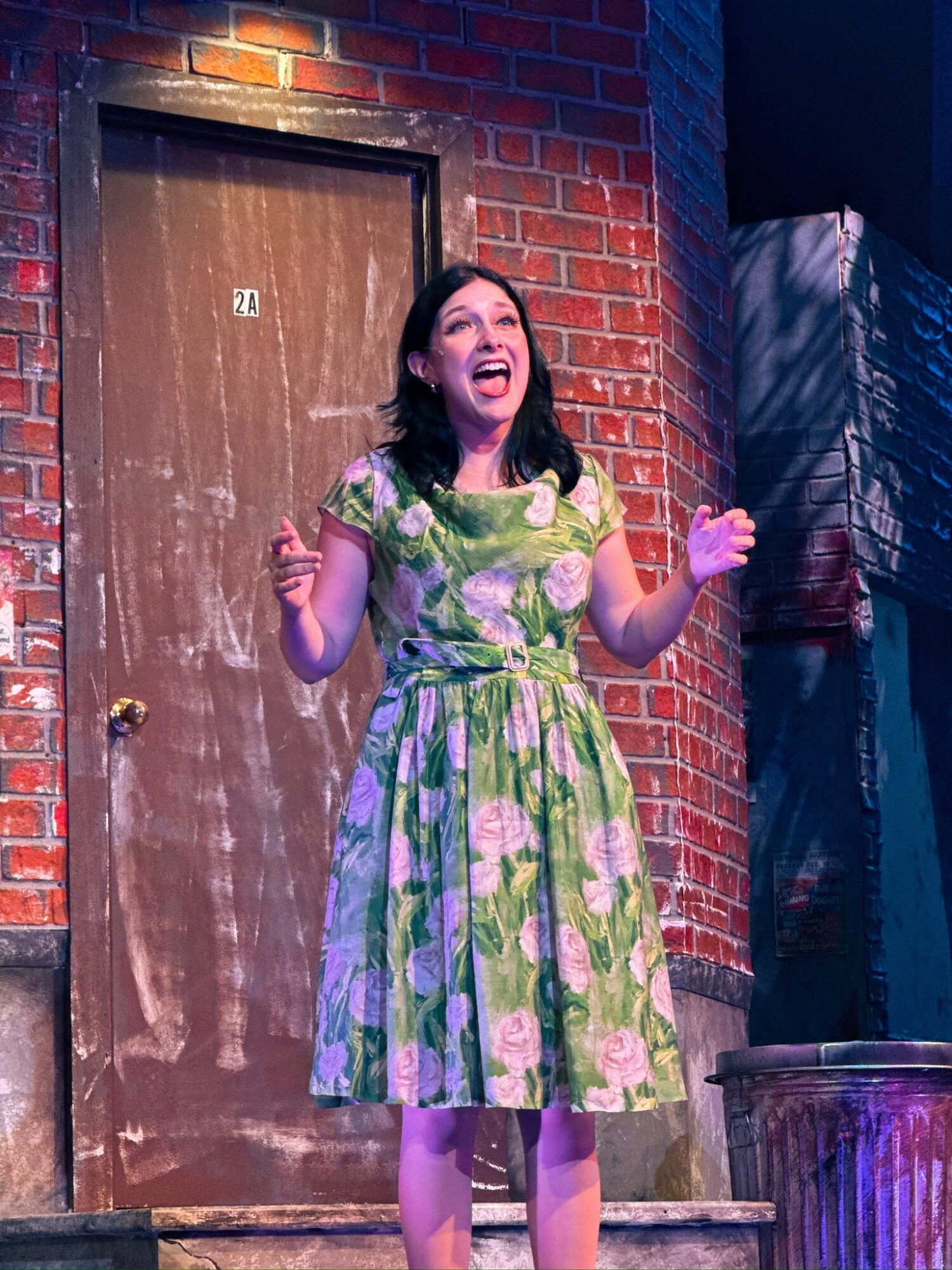 Woman in a floral dress smiling and gesturing on stage with brick wall background.