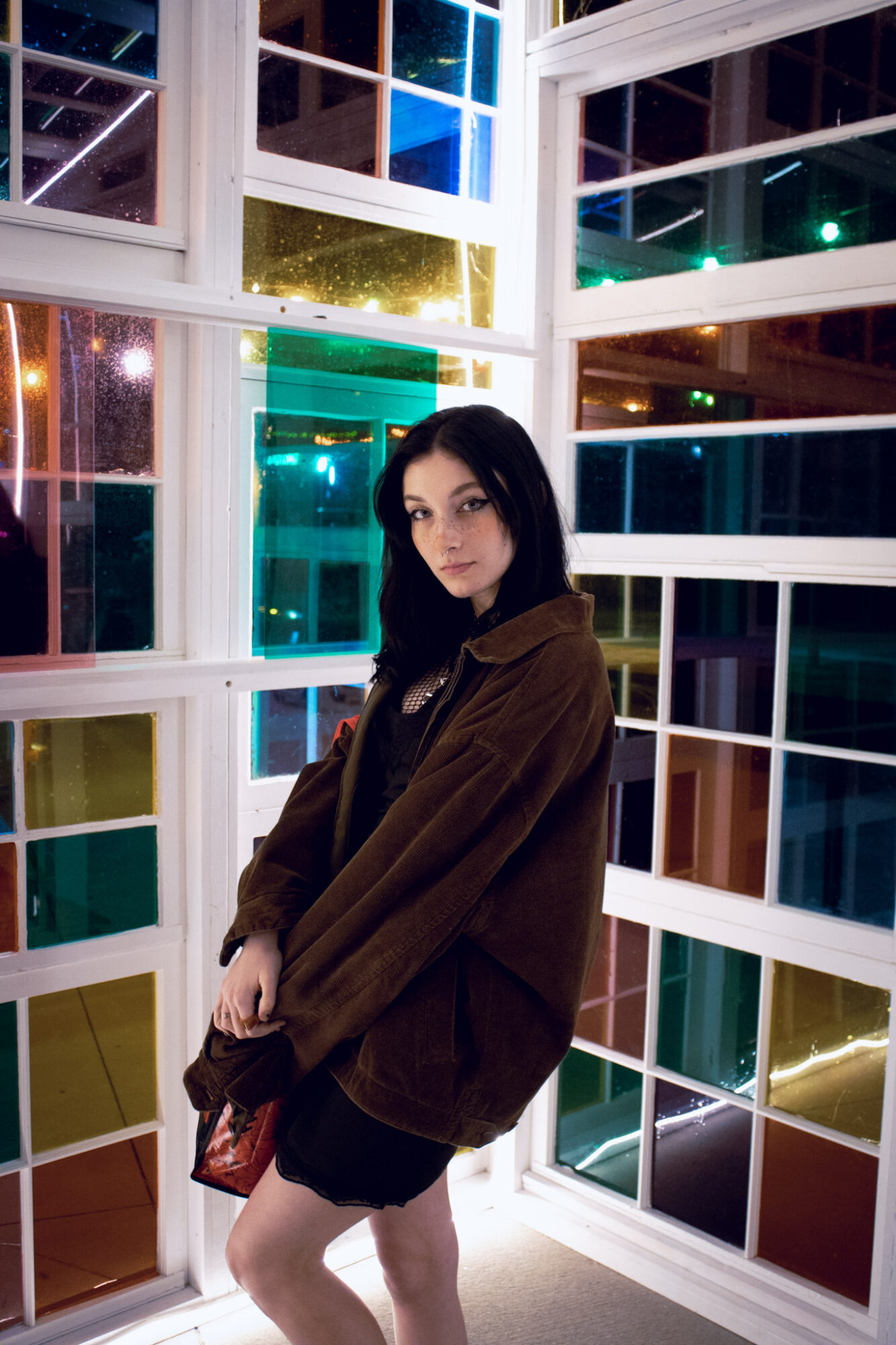 Young woman with dark hair in a brown jacket standing inside a colorful glass structure at night.
