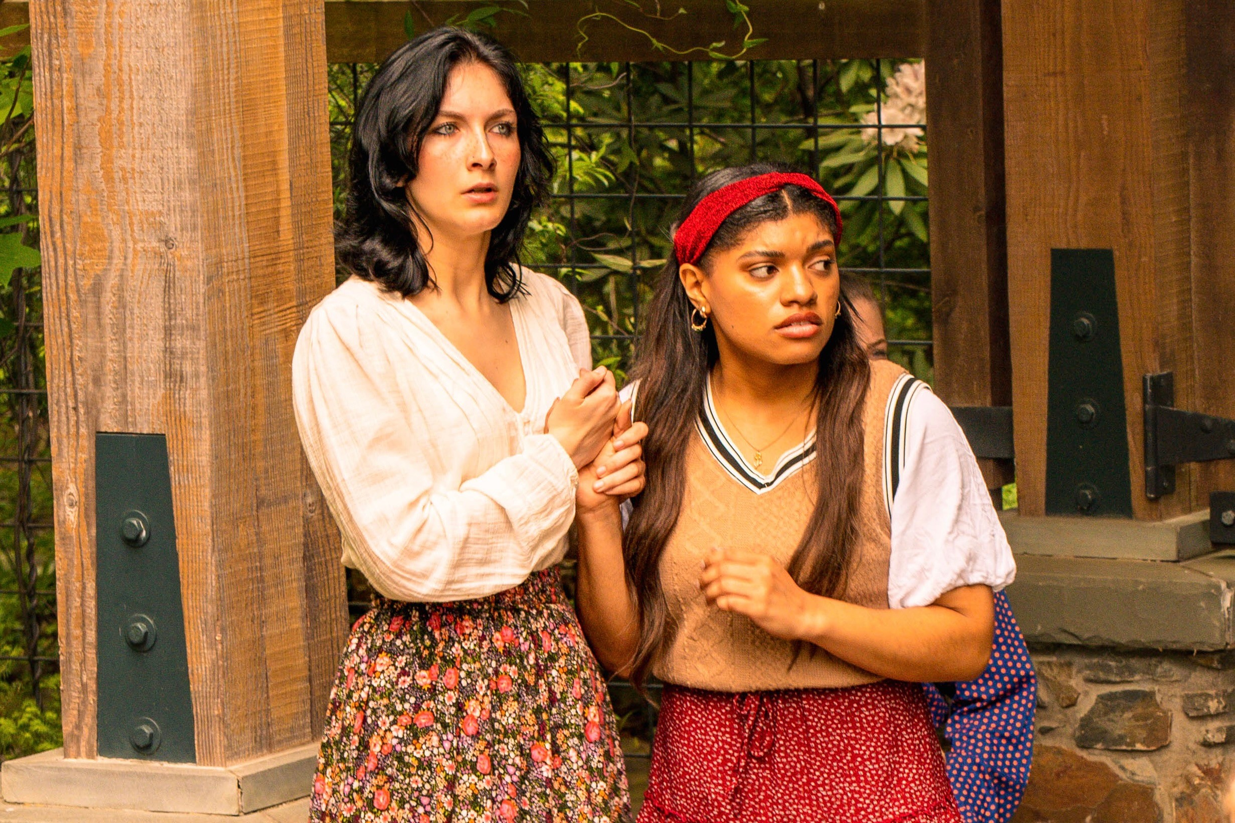 Two women standing indoors with wooden walls and greenery in the background, one with black hair and the other with brown hair and a red headband.