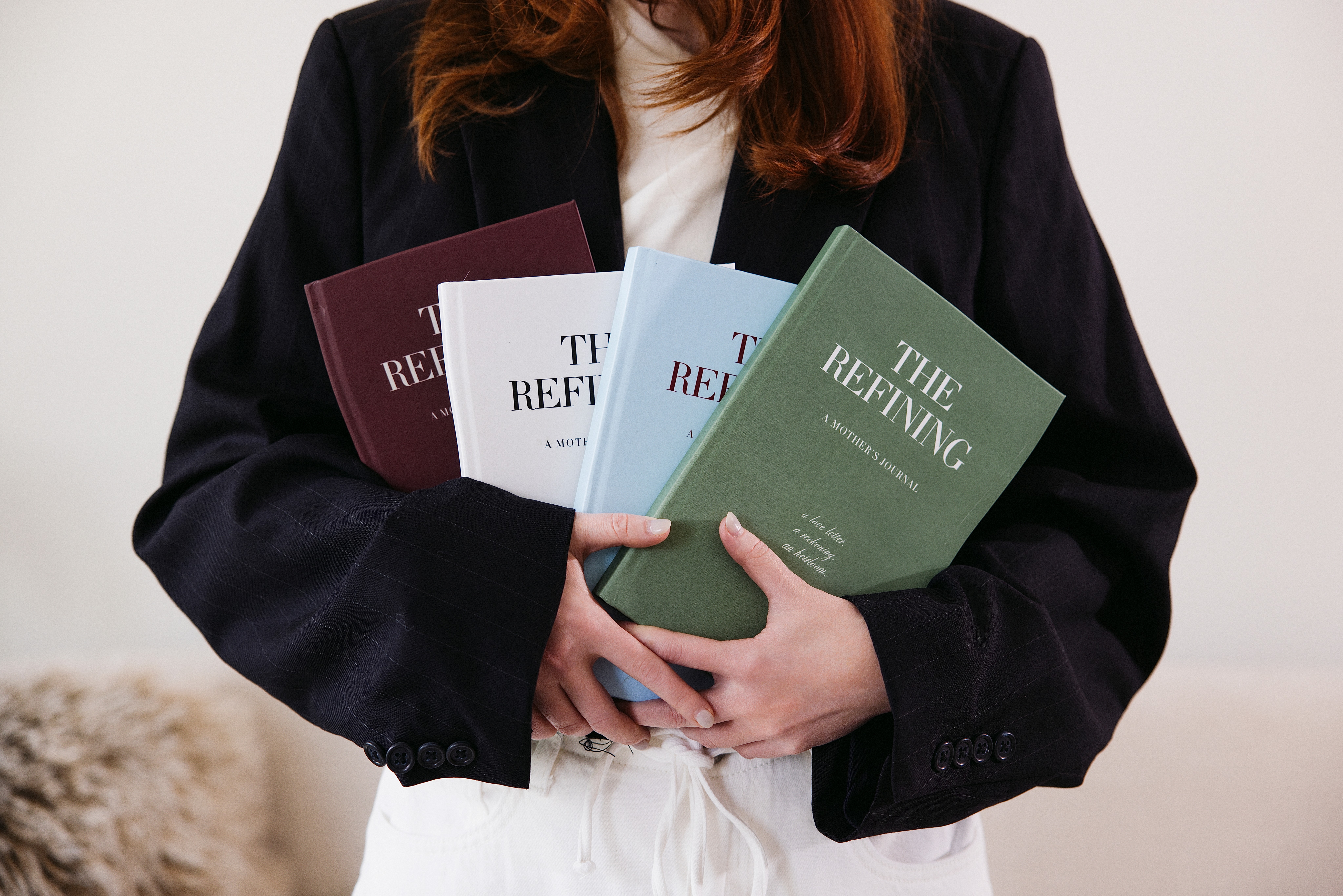 Person holding four books titled 'The Refining' in various colors, wearing a black blazer and white shirt, against a plain background.
