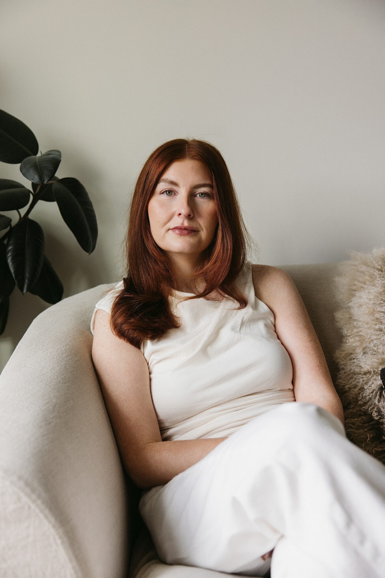 Woman with long brown hair sitting on a light-colored sofa, wearing a white sleeveless top, with a plant and cushion nearby.