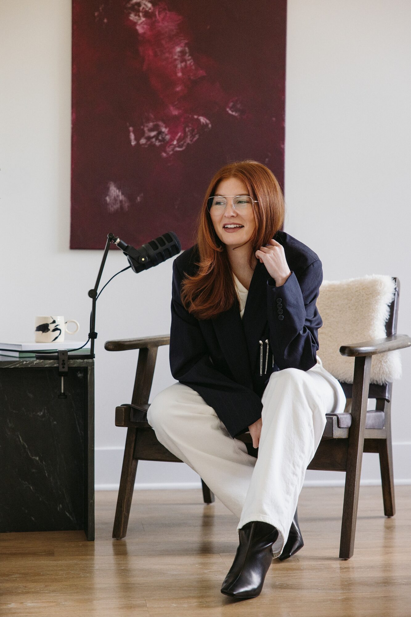 Woman with red hair smiling, sitting on a wooden chair, in a room with a large abstract painting on the wall.